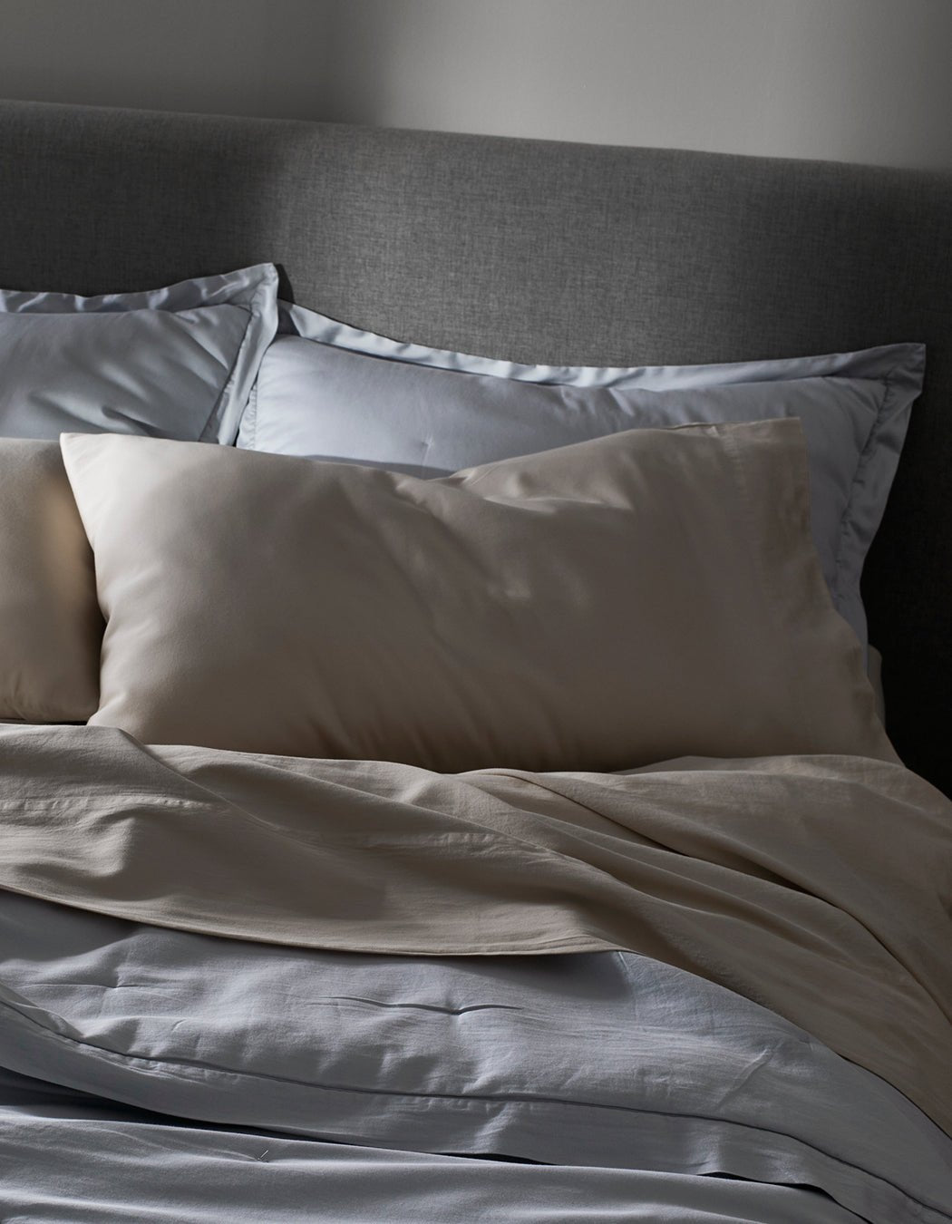 A softly lit room features a neatly made bed with Cozy Earth Soft-Wash Cotton Pillowcases in light gray and beige, paired with a plush comforter and set against a gray upholstered headboard.