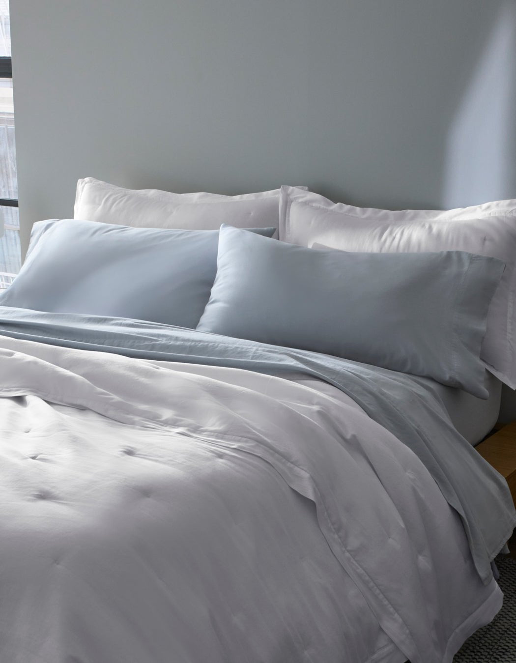 A bed near a window features Cozy Earth Soft-Wash Cotton Pillowcases in light gray and white, with matching bedding and two large pillows, all set against a pale gray wall.