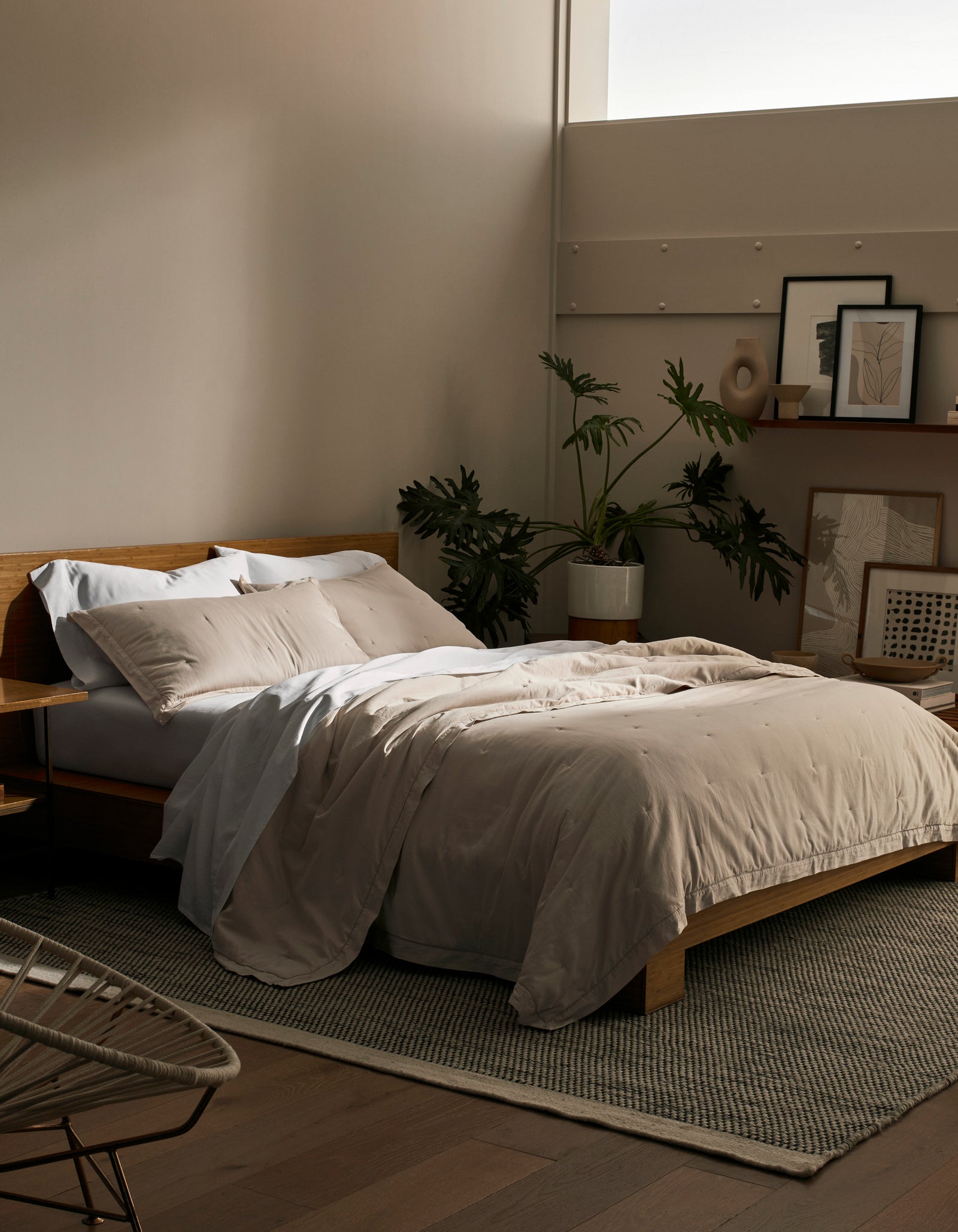 A cozy bedroom features a wooden bed frame with Cozy Earth's Soft-Wash Cotton Quilt in beige and white pillows. A large plant, framed artwork, and decor sit against the wall while soft natural light pours through a high window.