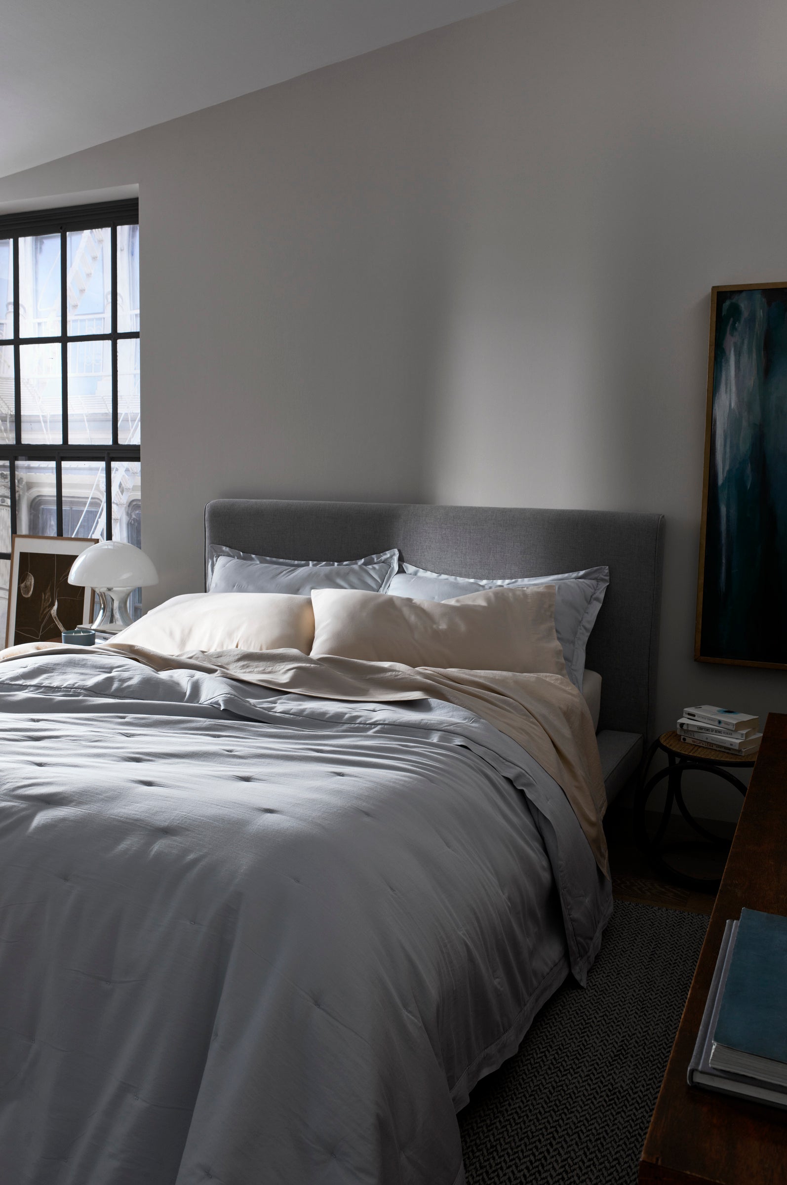 A neatly made bed features the Cozy Earth Soft-Wash Cotton Quilt in gray and cream, set in a softly lit bedroom with a large window, nightstand and lamp, wall painting, and books on a wooden dresser.