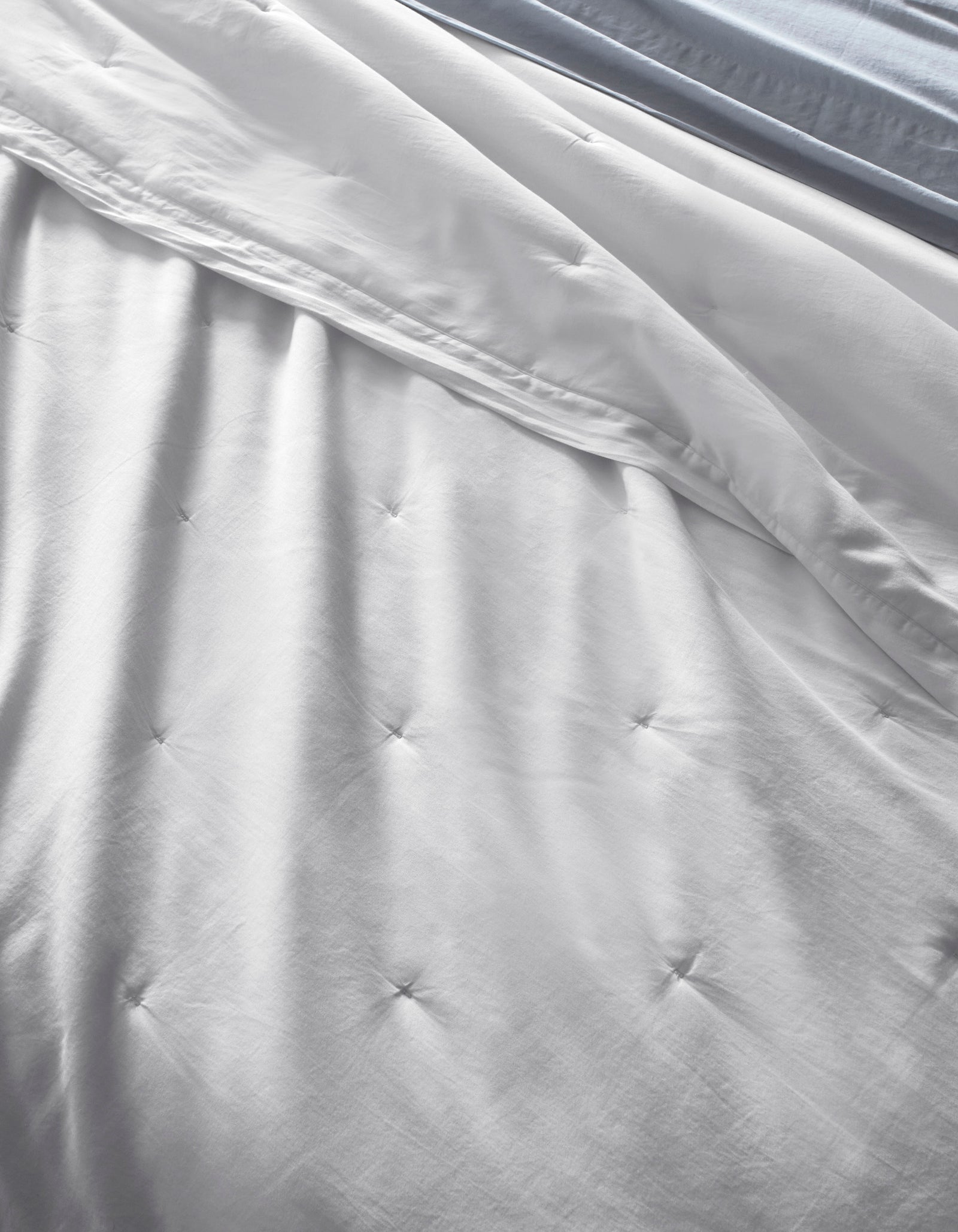 A close-up shows a neatly-made bed featuring the Cozy Earth Soft-Wash Cotton Quilt, white and tufted, with a light blue sheet peeking out. The smooth, soft fabric displays gentle folds and fine stitching for an inviting look.