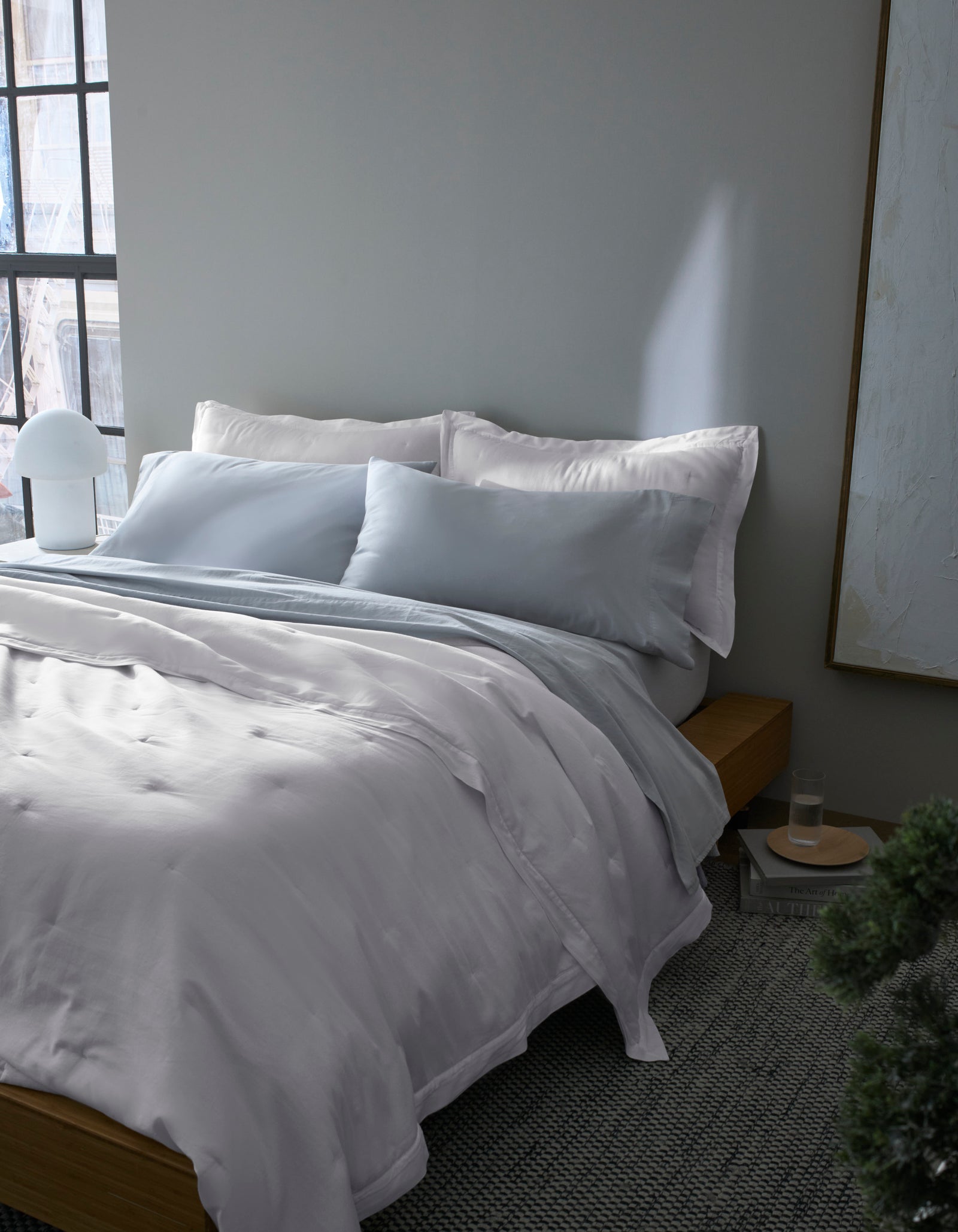 A neatly made bed with Cozy Earth's Soft-Wash Cotton Quilt in white and light gray sits by a window with soft natural light, next to a small round bedside table, lamp, and framed artwork. The room has calm, minimalist decor.