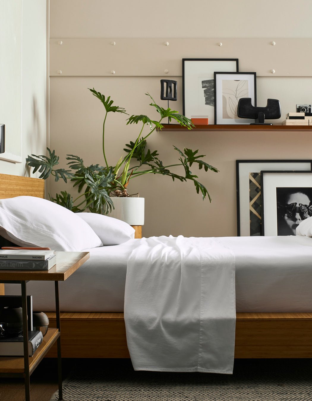 A modern bedroom features a wooden bed with Cozy Earth Soft-Wash Cotton Pillowcases, white bedding, and a potted plant on the bedside table. Art prints rest on a floating shelf by the beige wall, while a side table holds books.