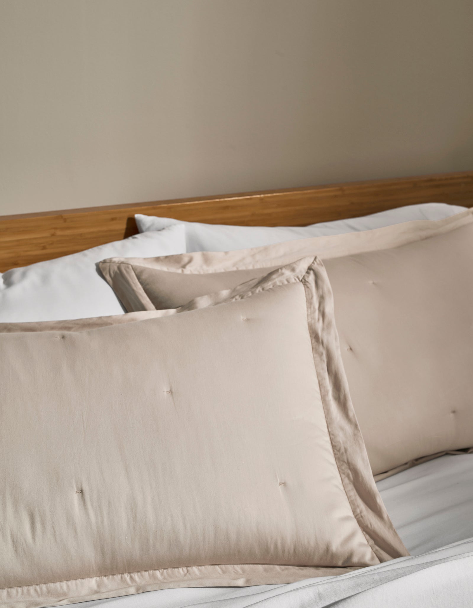 Two Cozy Earth Soft-Wash Cotton Quilted Shams in cream rest on a neatly made bed with white sheets and a wooden headboard against a beige wall.