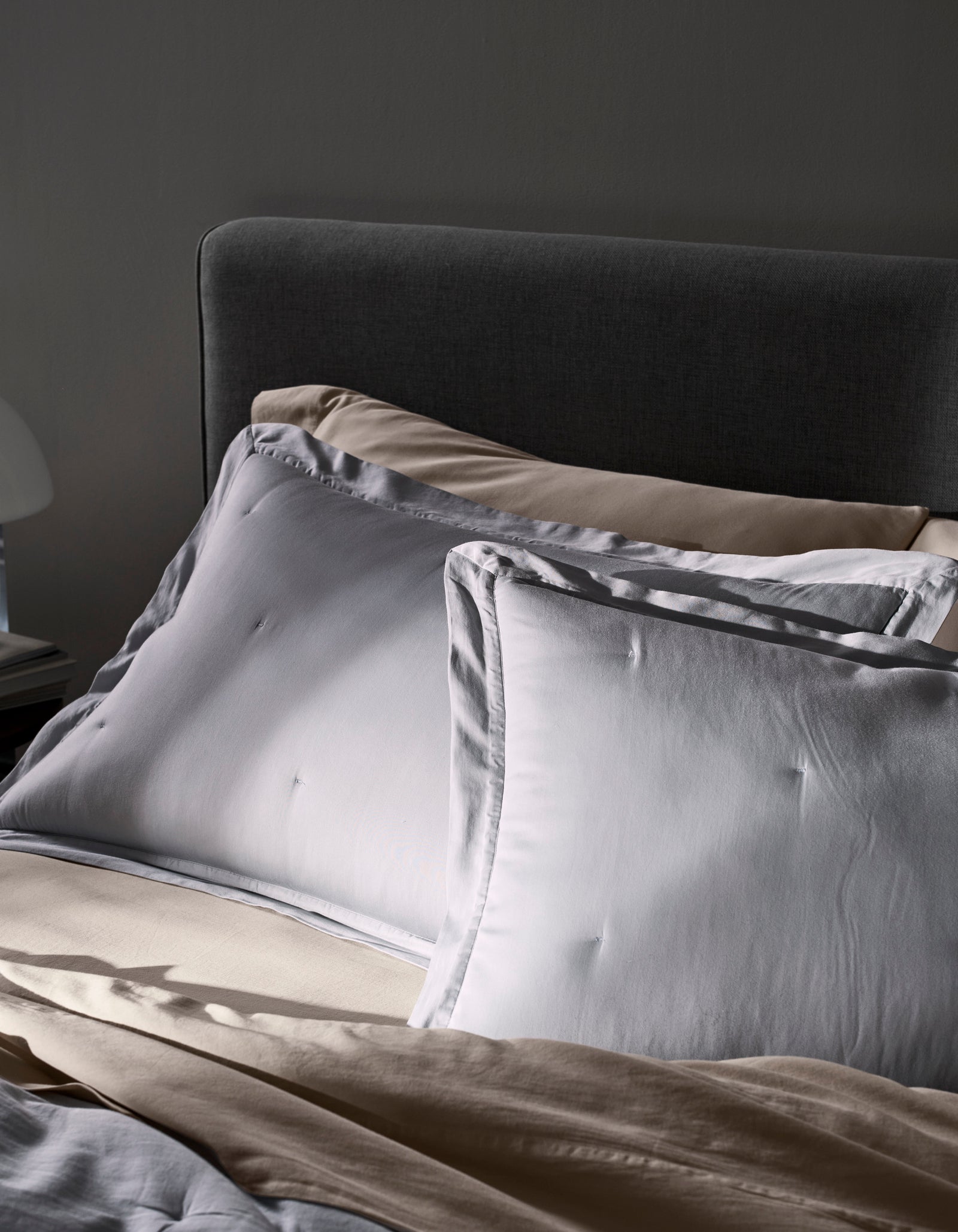 A close-up of an unmade bed features two Cozy Earth Soft-Wash Cotton Quilted Shams atop beige sheets and a dark gray upholstered headboard. Soft daylight highlights the calm, cozy atmosphere.