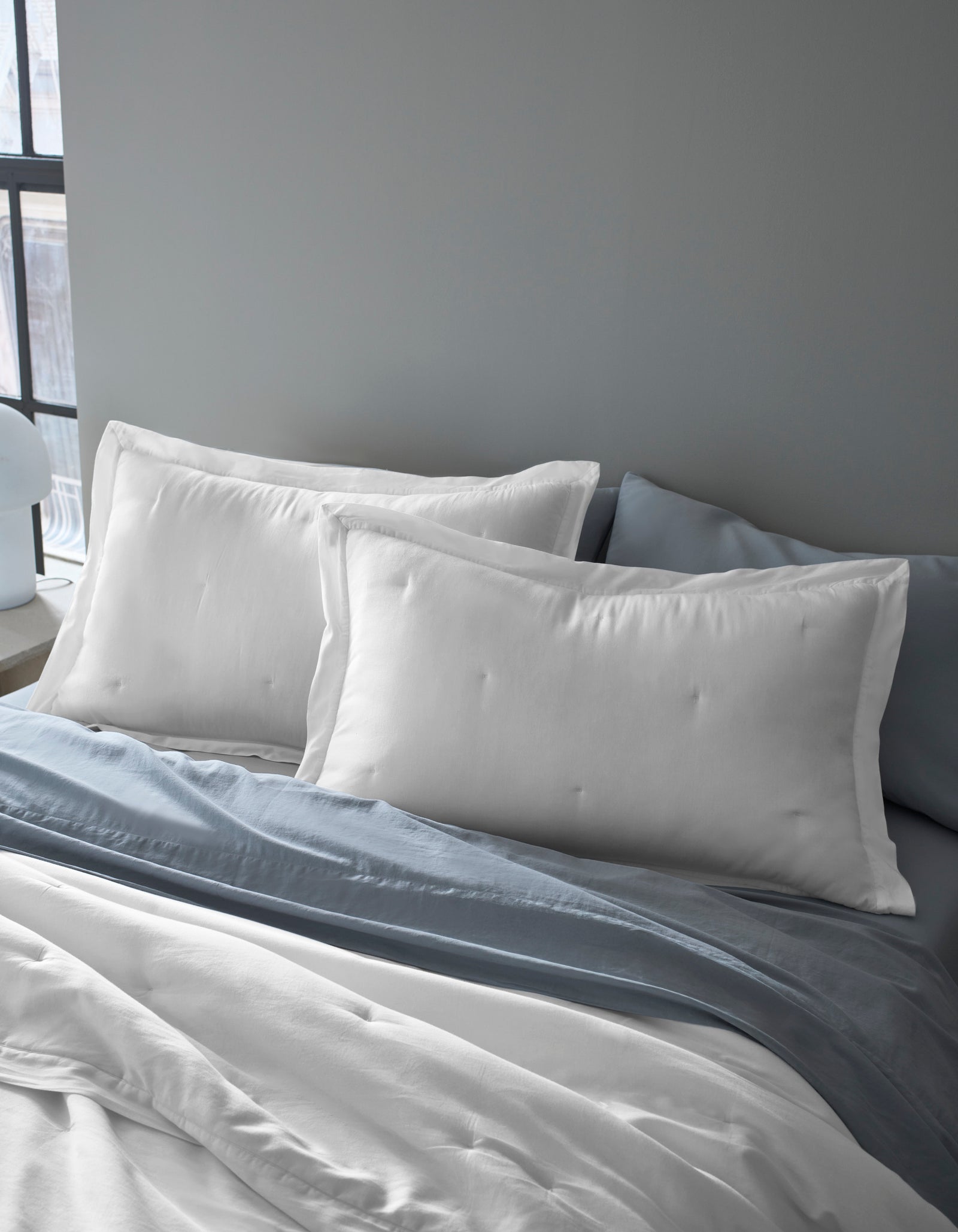 Two Cozy Earth Soft-Wash Cotton Quilted Shams are propped against a gray headboard on a bed with white sheets and a light blue-gray blanket in a softly lit room.