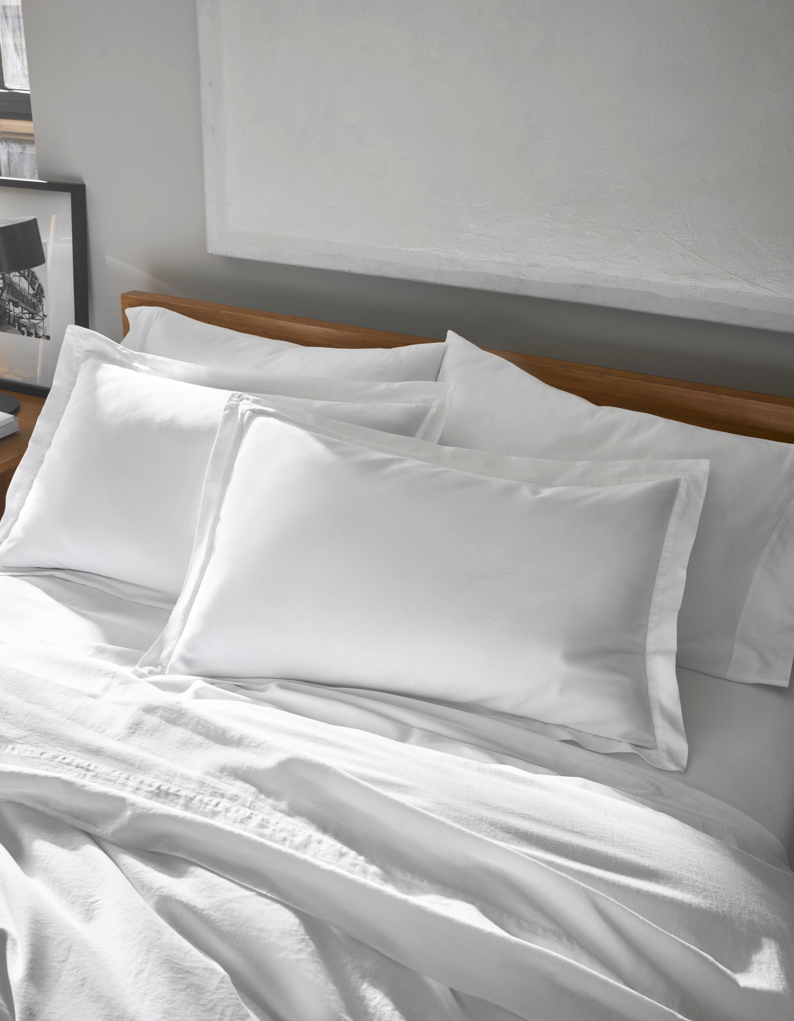 A neatly made bed features Cozy Earth's Soft-Wash Cotton Shams, crisp and white, paired with pillowcases against a wooden headboard in a softly lit, minimalist bedroom with a framed picture on the side table.