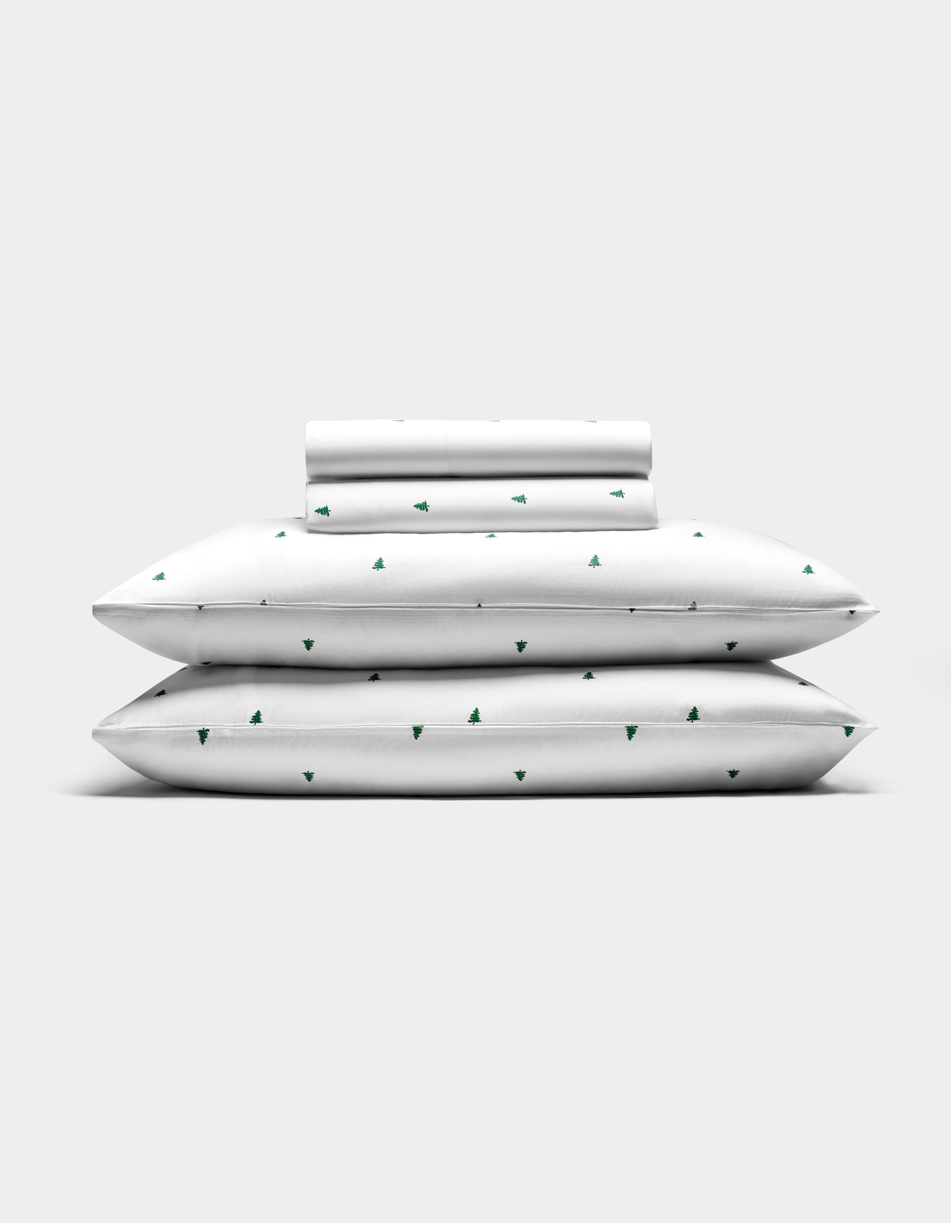 The Cozy Earth Bamboo Sheet Set features two pillows, a folded sheet, and a rolled pillowcase—all white with small green tree patterns—arranged against a plain light gray background.|Color:Jade Tree Snow