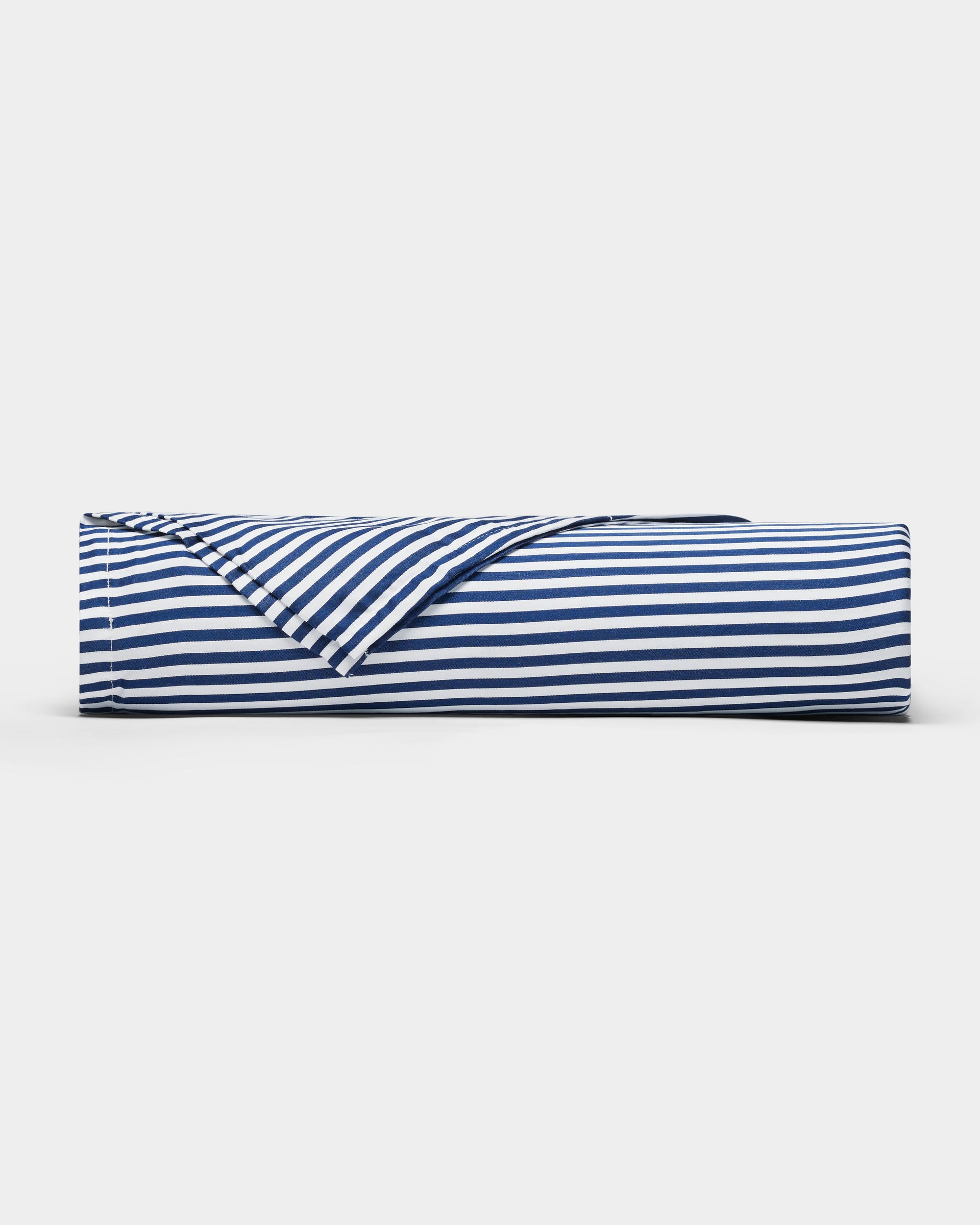 A Bamboo Sheet Set by Cozy Earth in navy blue and white stripes lies rolled on a plain white surface, with one corner partially unfolded to reveal the underside of the fabric.|Color:Classic Stripe in Pacific