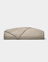 The Cozy Earth Bamboo Jersey Quilt, featuring a beige, quilted grid pattern, is neatly folded on a white background. A turned-down corner highlights its soft, luxurious texture.|Color:Driftwood