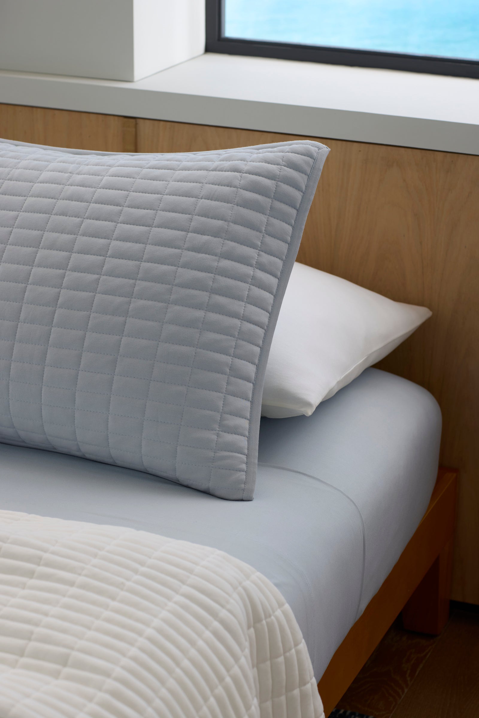 A bed featuring the Cozy Earth Bamboo Jersey Quilt Bundle—a light blue quilt and matching fitted sheet, layered over a white pillow and textured blanket, set against a wooden headboard and window.