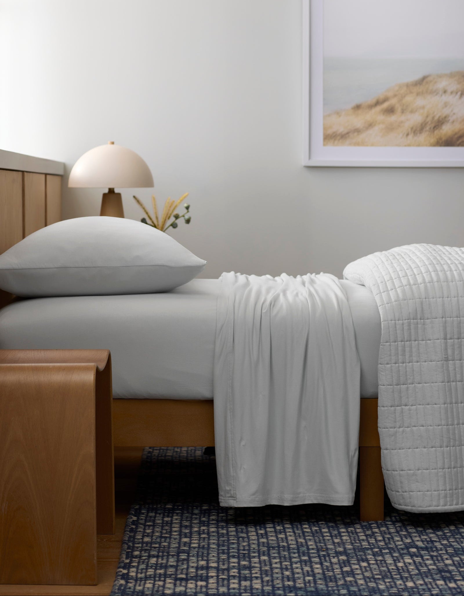 A bed made with Cozy Earth’s Bamboo Jersey Sheet Set rests against a wooden headboard. A wooden bench stands at the foot, while a lamp, small vase, and framed landscape photo accentuate the minimal, serene bedroom.