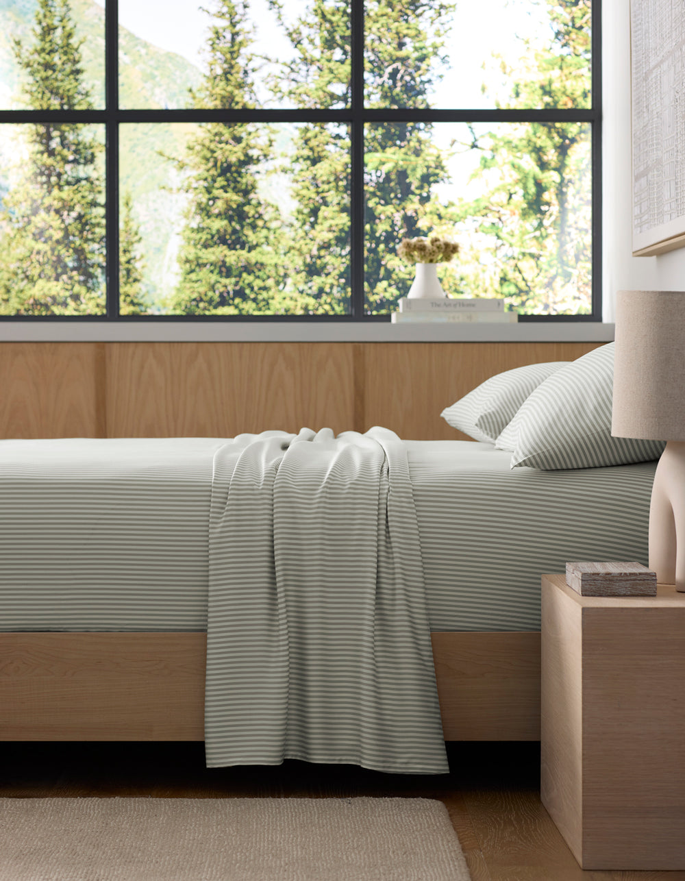 A neatly made bed features the Cozy Earth Bamboo Sheet Set in light gray stripes, paired with matching pillows and a throw, next to a wooden nightstand in a modern bedroom with large windows overlooking green trees.|Color:Classic Stripe in Laurel