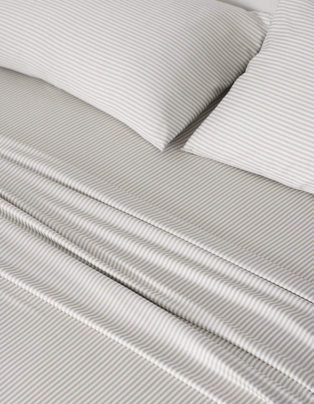A close-up of a neatly made bed features the Cozy Earth Bamboo Sheet Set—light gray and white thin stripes on sheets and two pillows—offering a soft, orderly look.|Color:Classic Stripe in Laurel