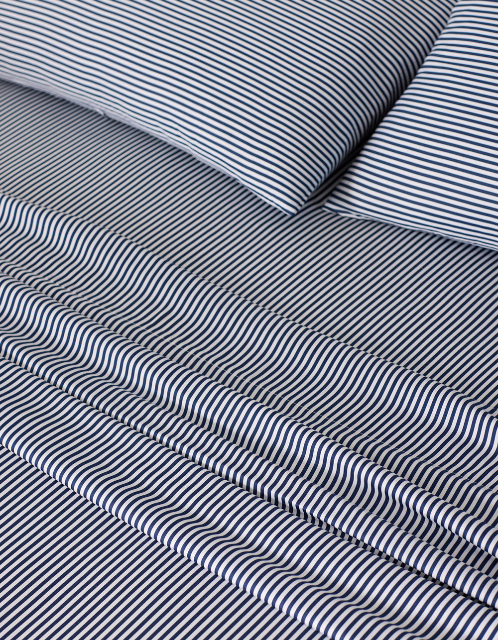 A close-up of a neatly made bed showcases the Cozy Earth Bamboo Sheet Set, featuring a smooth, white and dark blue narrow pinstripe pattern on the pillow and sheets.|Color:Classic Stripe in Pacific