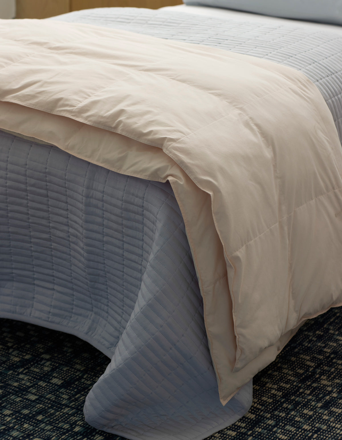 A neatly made bed features a light gray quilted coverlet with the Cozy Earth Down Alternative Throw, a pale beige comforter, folded at the foot, all set on a dark patterned carpet.