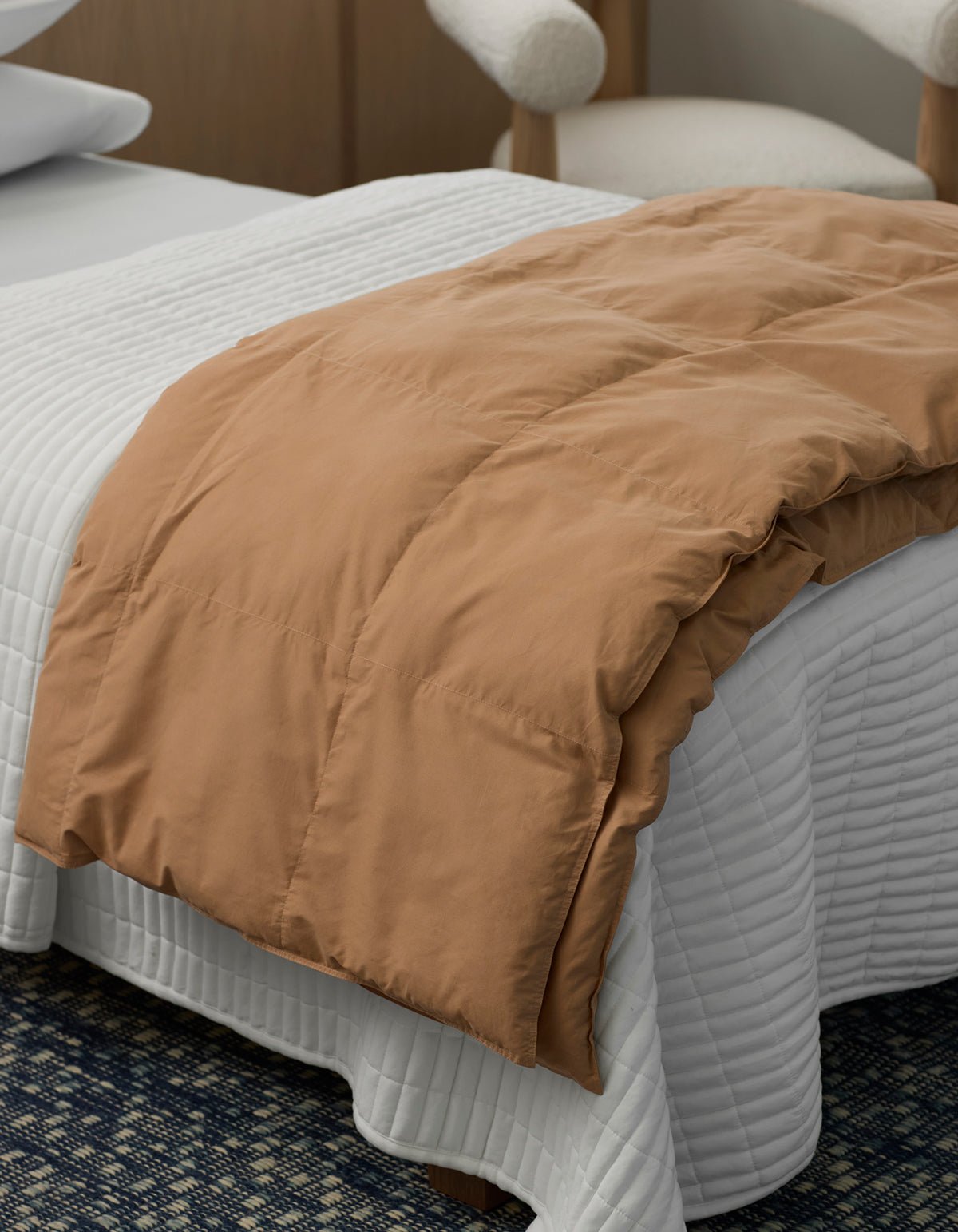A Cozy Earth Down Alternative Throw in tan is neatly draped over the foot of a bed with a white quilted bedspread, in a modern room featuring a cushioned chair in the background.