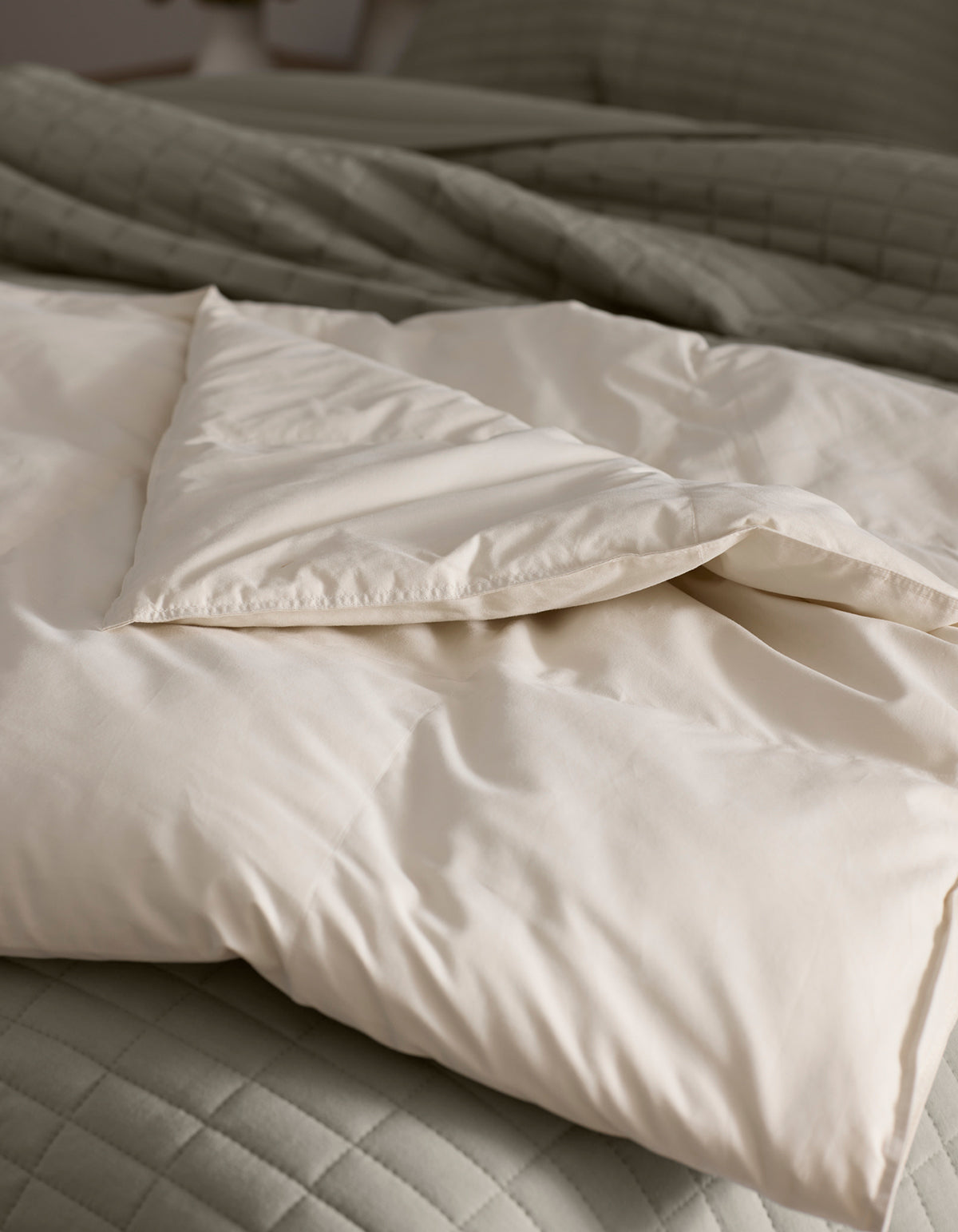 A close-up of the Cozy Earth Down Alternative Throw, cream-colored and partially folded back, resting on a textured light gray quilted bedspread.