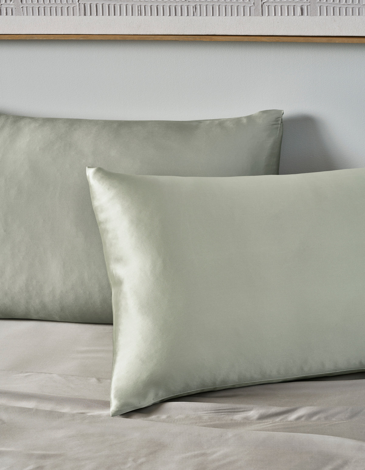 Two Cozy Earth Silk Pillowcases in pale green rest on a matching sheet, set against a light gray wall with part of a wooden frame visible above.