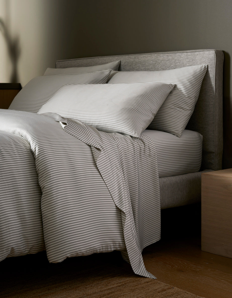 A neatly made bed with Cozy Earth's Bamboo Bedding Deluxe Bundle - Silk in light gray and white stripes sits beside a light wooden nightstand in a softly lit, minimalistic bedroom.