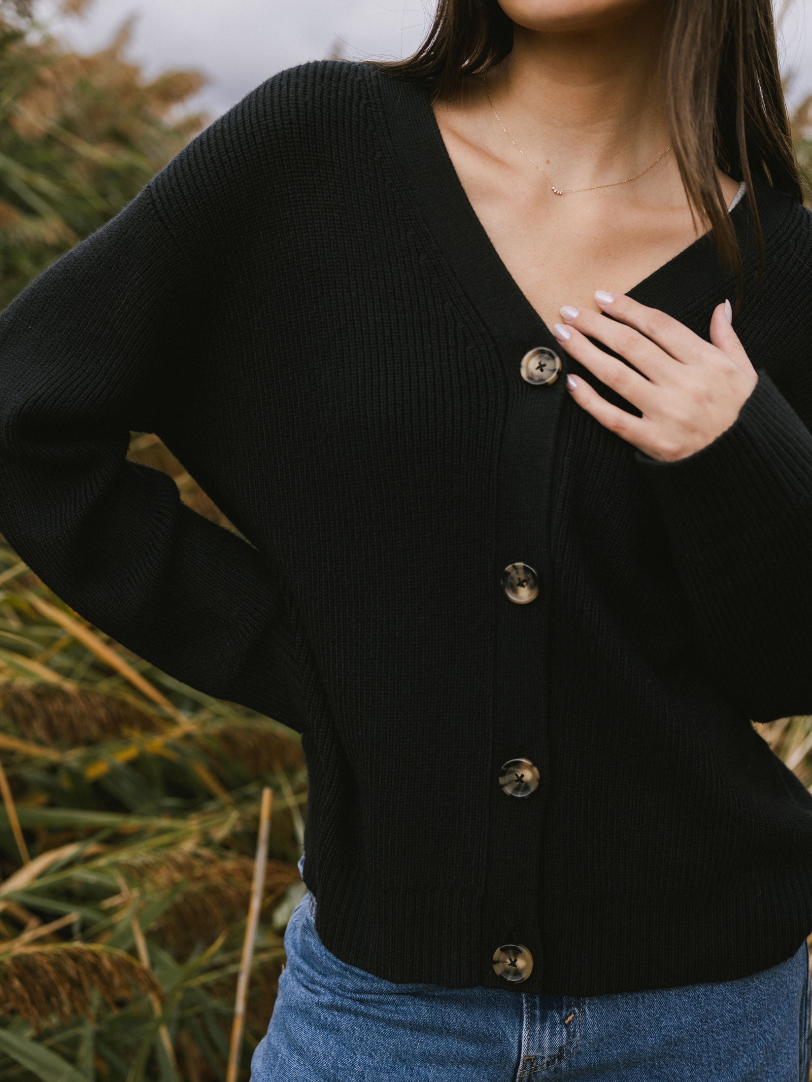 A woman stands surrounded by tall grass wearing Cozy Earth Classic Cardigan in Black. She is shown from the neck down and waist up