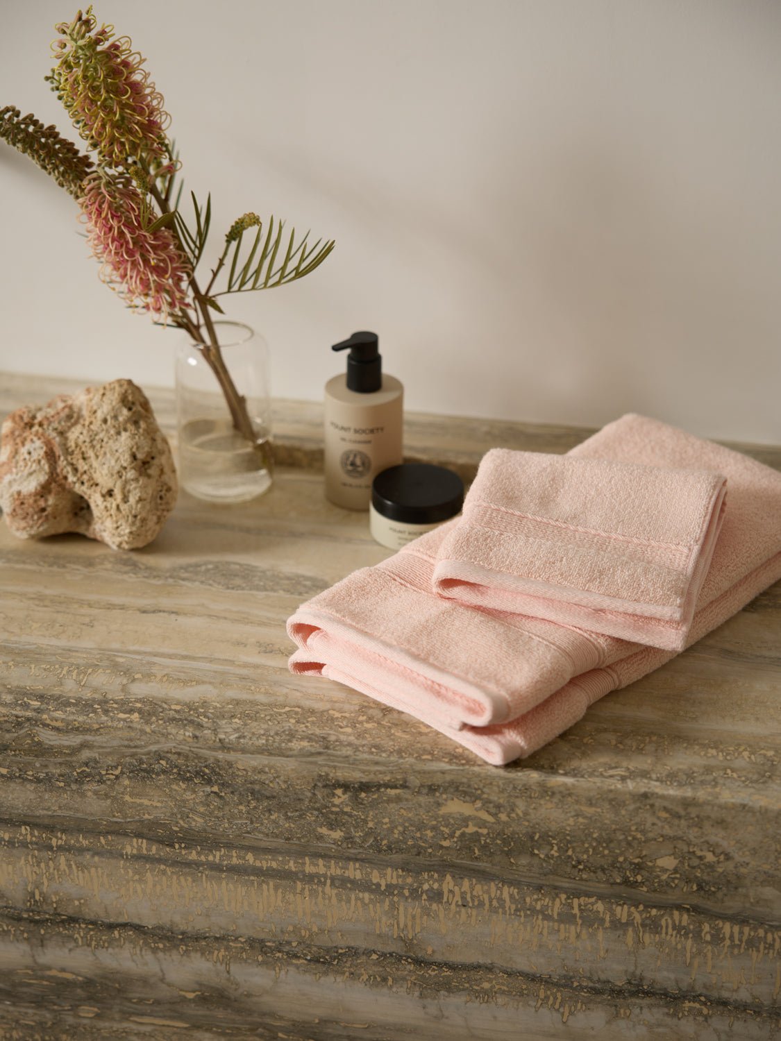 A neatly folded Luxe Washcloth set by Cozy Earth rests on a marble countertop. Beside it are a natural sponge, a small potted plant with red spiky flowers, and two bottles of skincare products, creating a spa-like atmosphere.
