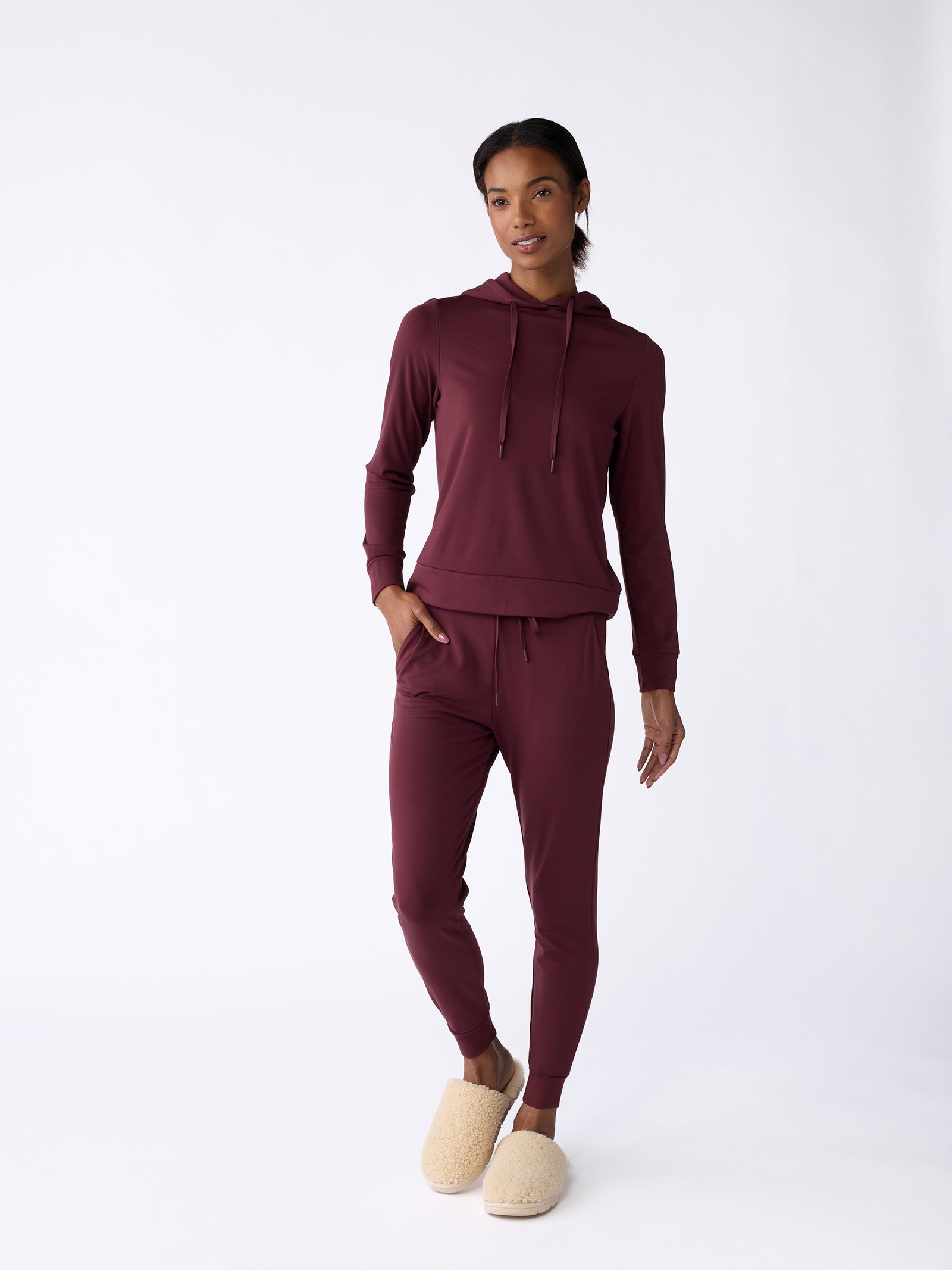 A person wearing Cozy Earth's maroon Women’s Bamboo Jogger Pant and a matching hoodie stands against a plain white background. They have their hands in their pockets and are wearing beige slippers.