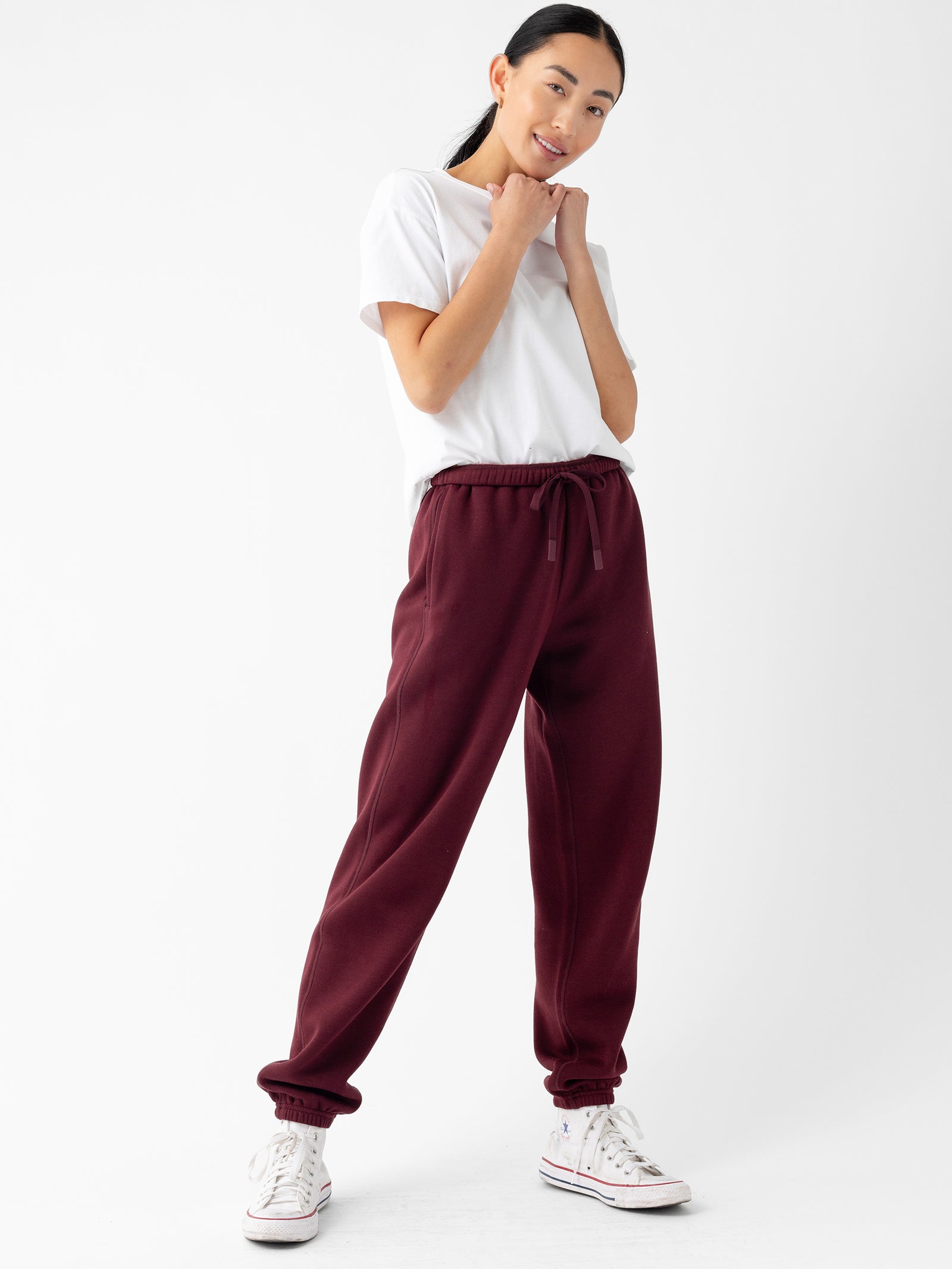 A person stands against a plain white background wearing a white T-shirt, Cozy Earth's Women's CityScape Sweatpant in maroon, and white sneakers with red and blue stripes. They have their hands near their face and are smiling, conveying a relaxed and cheerful demeanor.