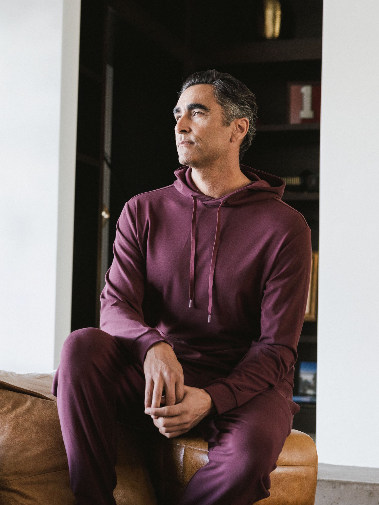 Burgundy Bamboo Hoodie worn by man in a living room of a house. The man is sitting on the arm of a couch.