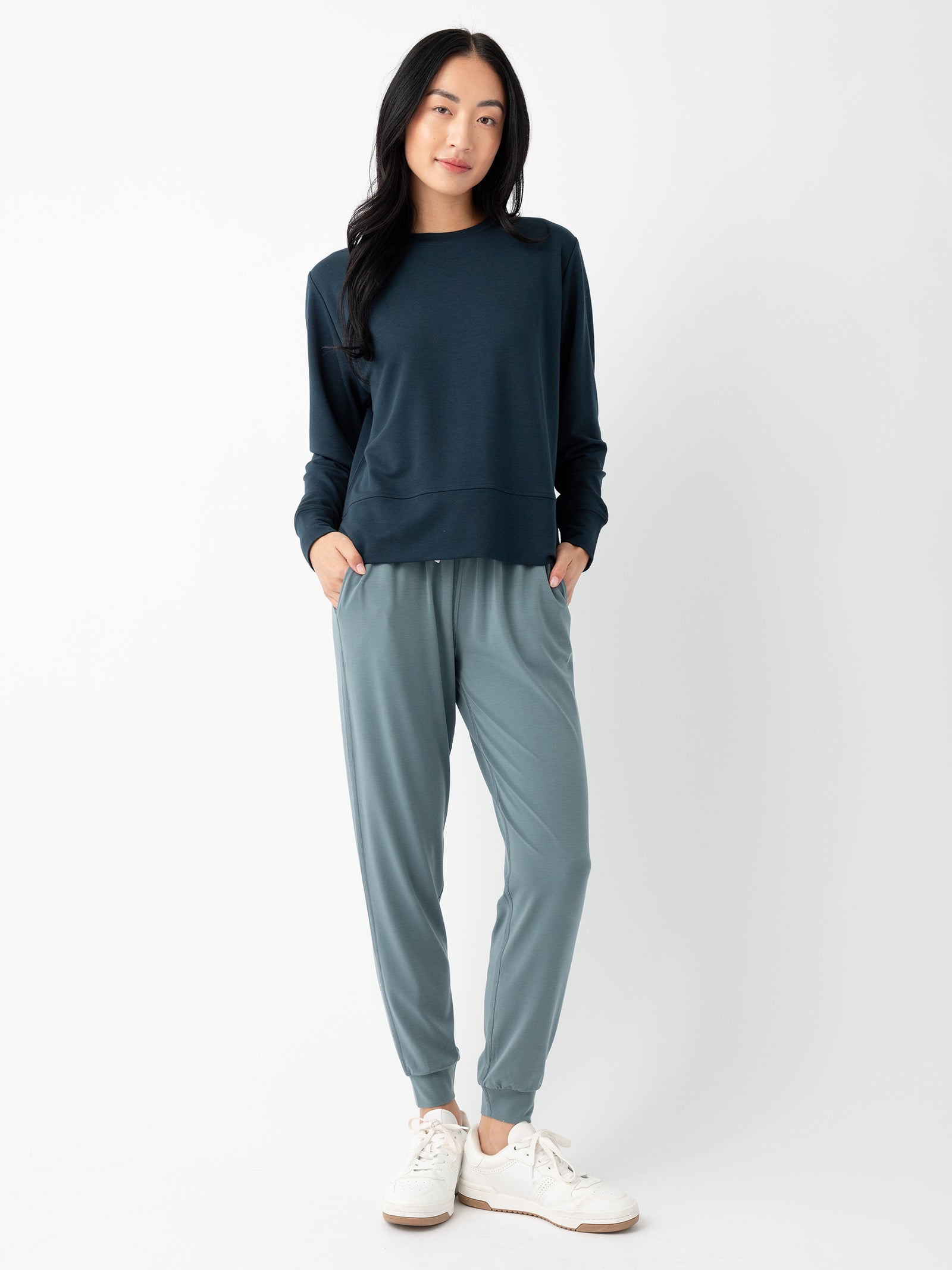 A person with long black hair, wearing a dark blue long-sleeve shirt and light blue Women's Studio Jogger pants by Cozy Earth, is standing against a plain white background. Their hands are tucked into the pockets of the pants, and they are wearing white sneakers.
