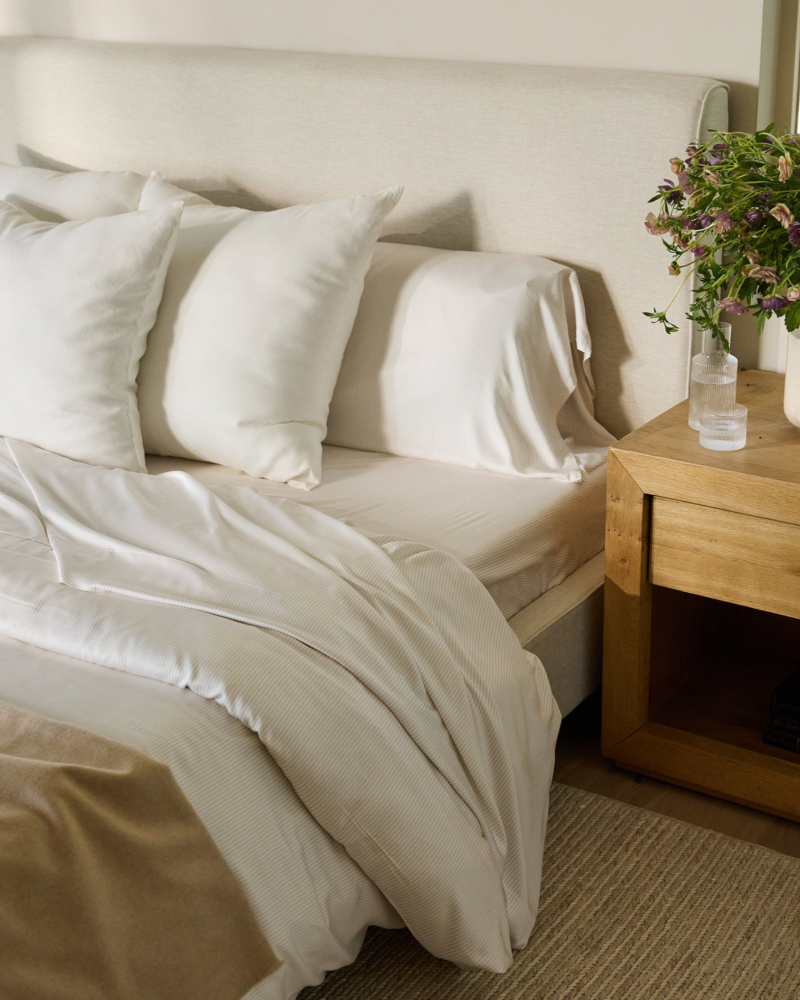 A cozy bedroom scene featuring a bed adorned with Cozy Earth's Bamboo Duvet Cover, complemented by white pillows and sheets, a beige blanket, and a light beige headboard. A wooden nightstand holds a potted plant and a glass of water, with books neatly stacked below. A window with greenery is in the background.