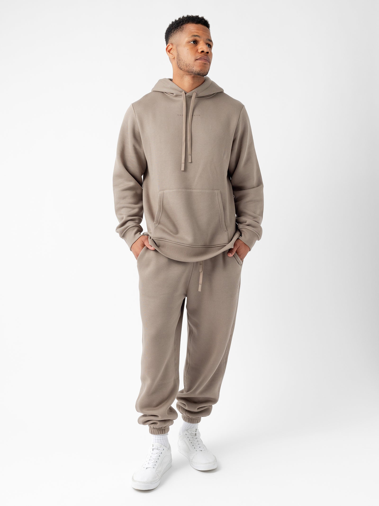 A man stands against a plain white background wearing the taupe-colored Men's CityScape Hoodie and matching jogger pants by Cozy Earth, paired with white high-top sneakers. He has his hands in the hoodie pocket and is looking off to the side.