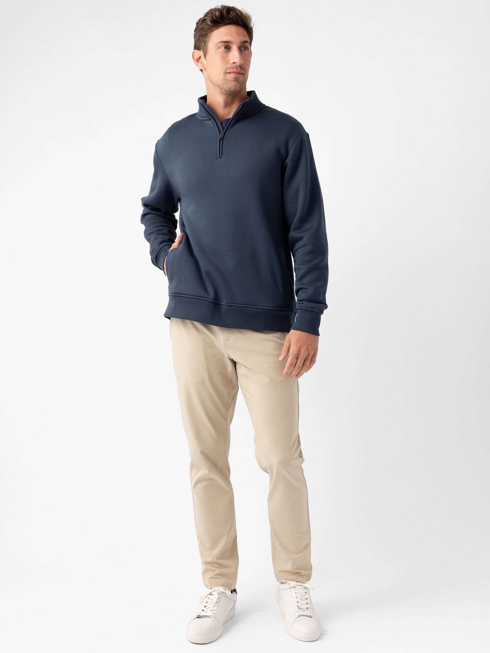 A man stands in a relaxed pose against a white background. He is wearing a dark blue Cozy Earth Men's CityScape Quarter Zip, light tan pants, and white sneakers. One hand is in his pocket while the other hangs down by his side.