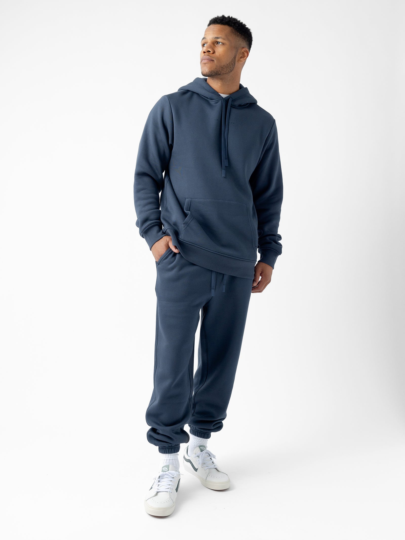 A man stands against a plain white background wearing a dark blue hoodie and Cozy Earth's Men's CityScape Sweatpant. His hands rest in the pockets of the hoodie, and he pairs the outfit with white sneakers. He gazes to the right with a slight smile.