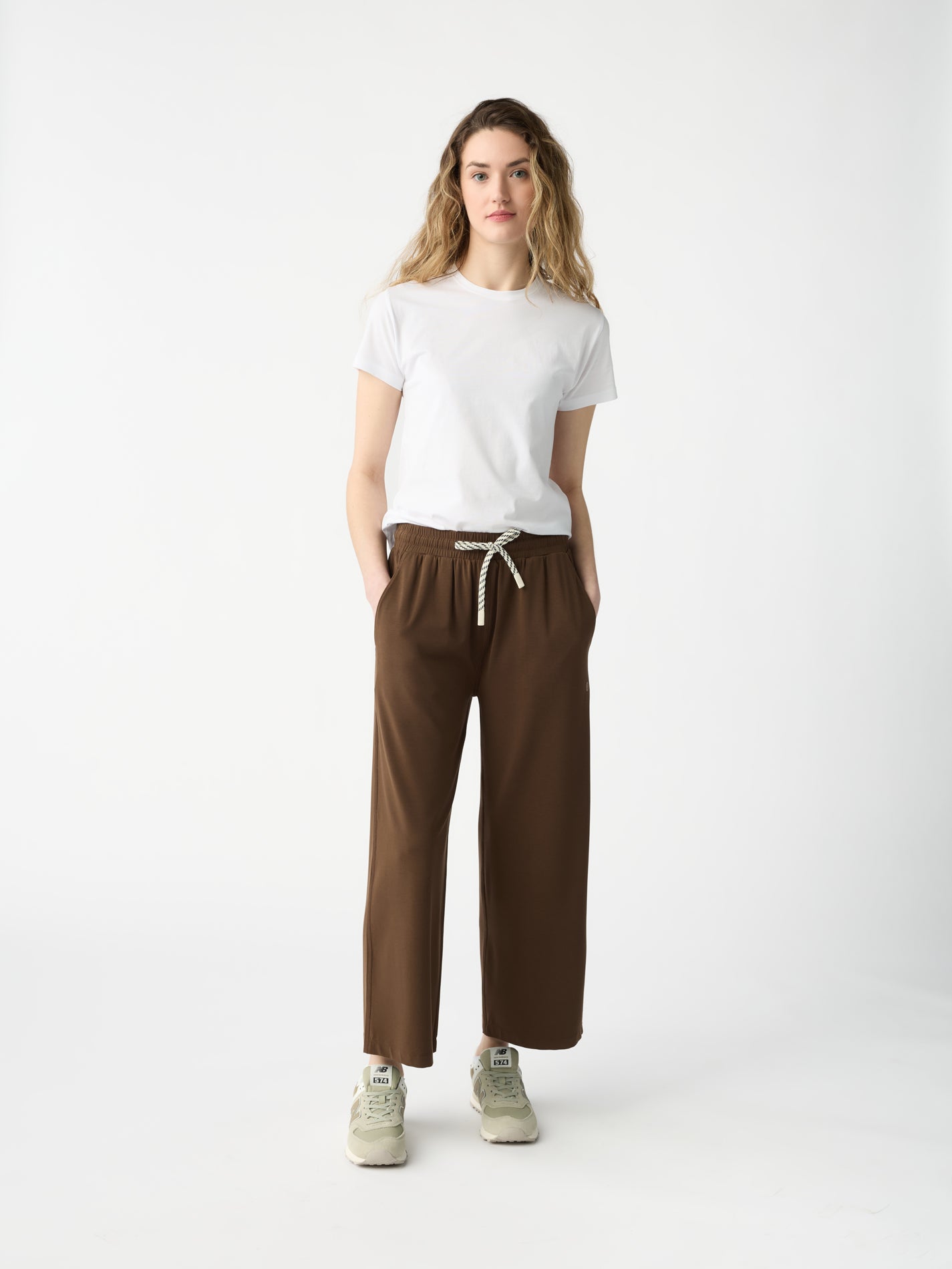 A person with long wavy hair stands against a white background, wearing a white T-shirt, Cozy Earth's Women's Studio Cropped Wide Leg Pant in brown with a drawstring, and beige sneakers. Their hands are in their pockets.
