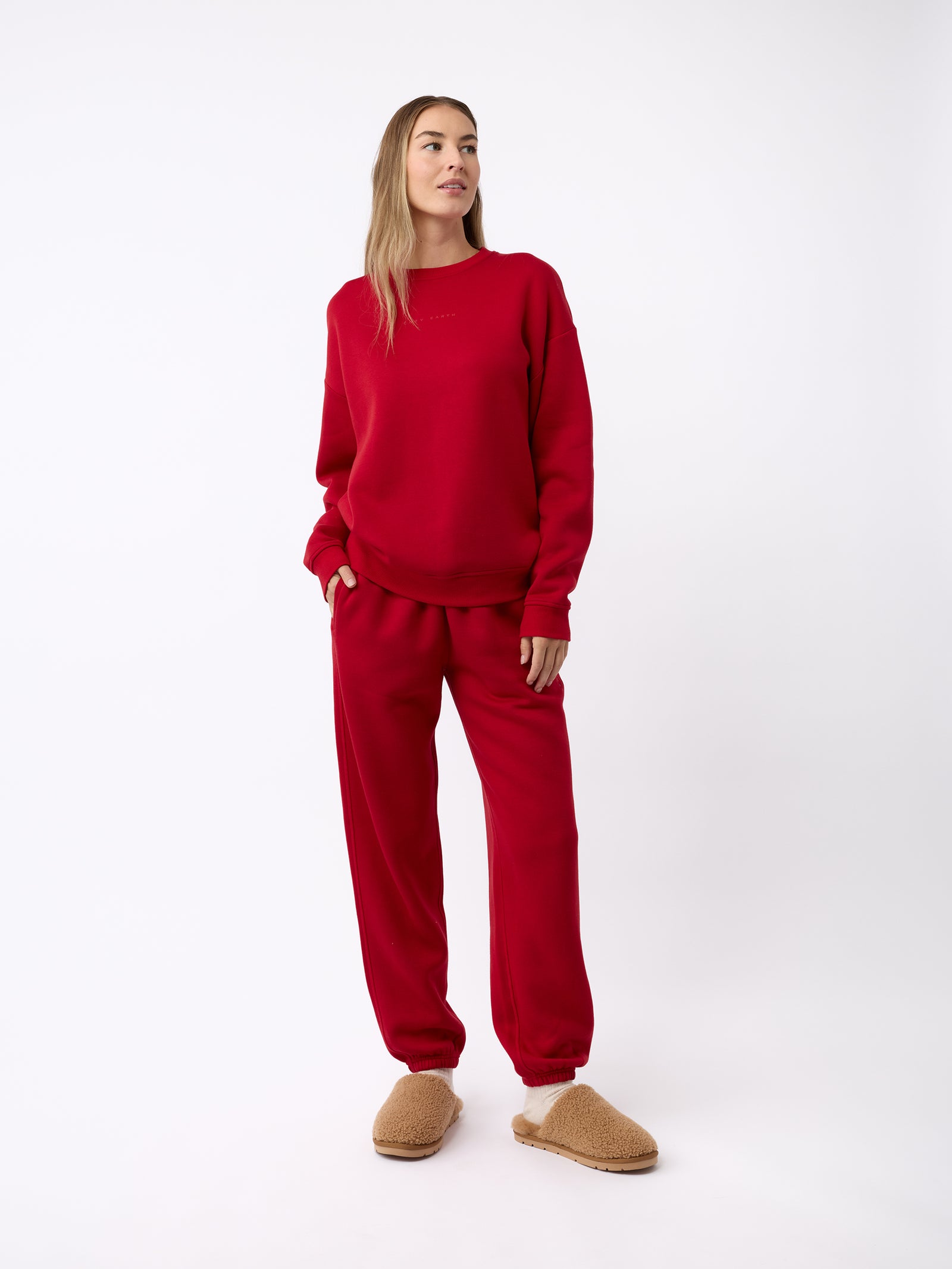 A person stands against a plain background, wearing a matching red sweatshirt and sweatpants set. They have light brown hair and are gazing to the side. On their feet, they are wearing Cozy Earth's Puffy Sheep Slippers in tan, which look fuzzy and comfortable. They have one hand in their pocket.