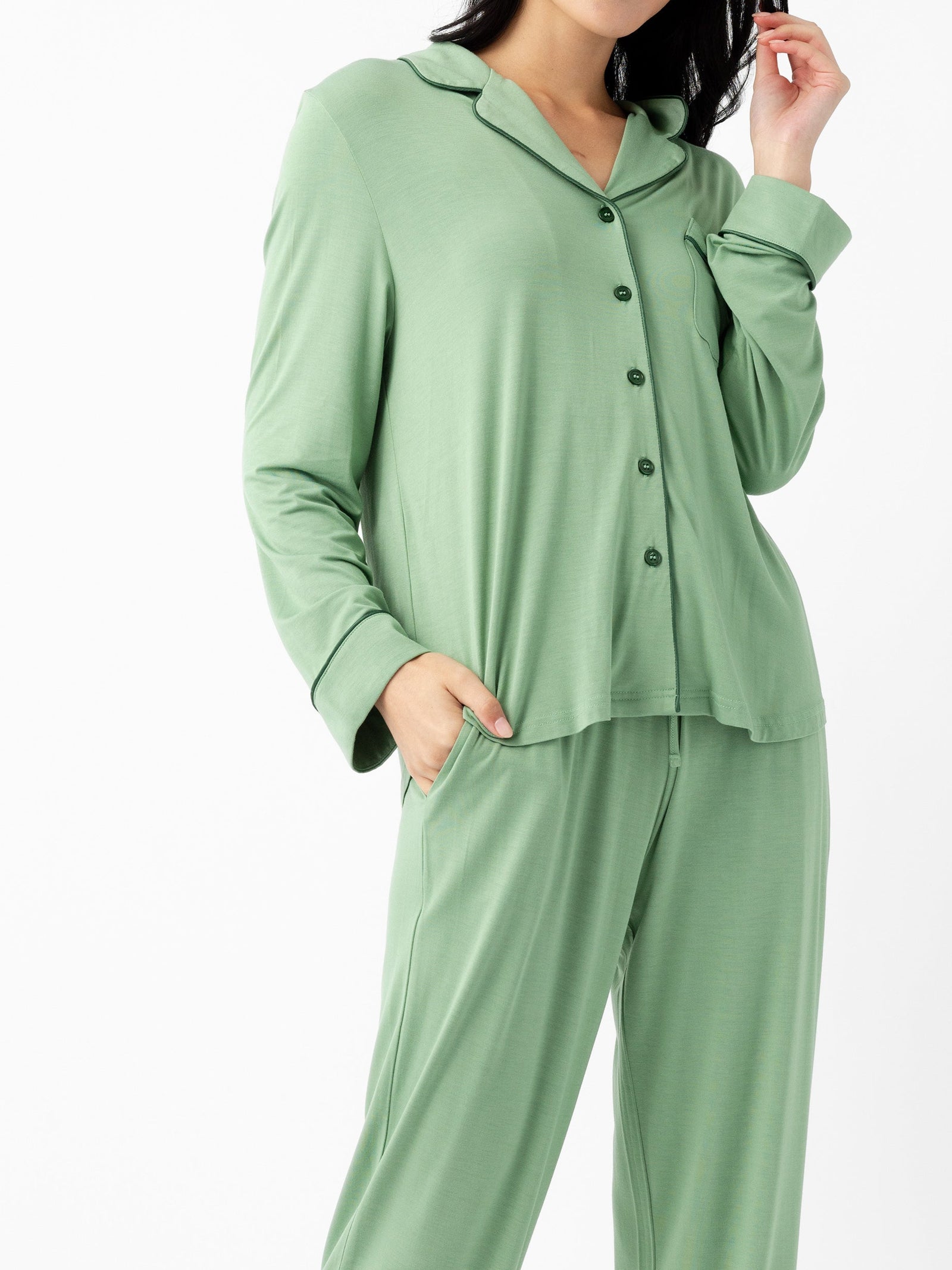 A woman wears the Cozy Earth Last Chance Women's Bamboo Stretch-Knit Long Sleeve Pajama Set in light green, featuring a loose fit, button-up top, and matching pants, with one hand in her pocket against a plain white background.