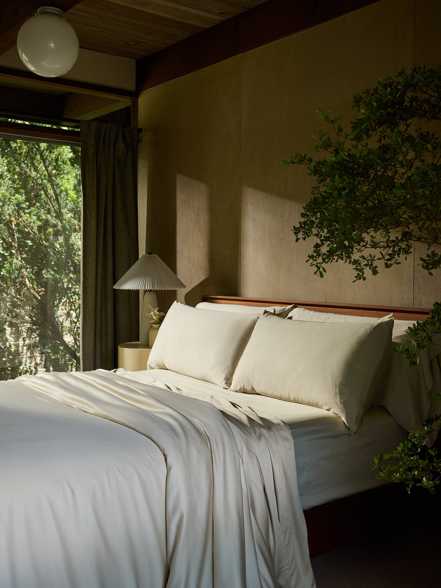 A serene bedroom with a large window showcasing lush greenery outside. The bed is made with Cozy Earth's Bamboo Flat Sheet and pristine white pillows, complemented by a beige headboard. A beige lamp with a pleated shade sits on a nightstand, and natural light filters through the room.