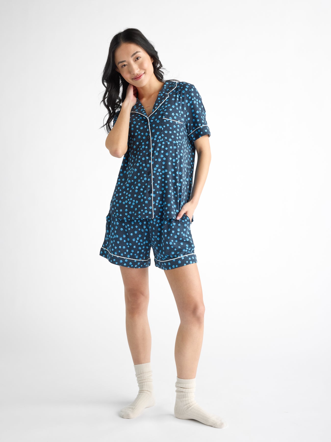 A person wearing a blue polka dot, Women's Bamboo Stretch-Knit Short Sleeve Pajama Top from HIDE with white trim smiles with one hand on their chin. They also have on white socks against a plain white background.