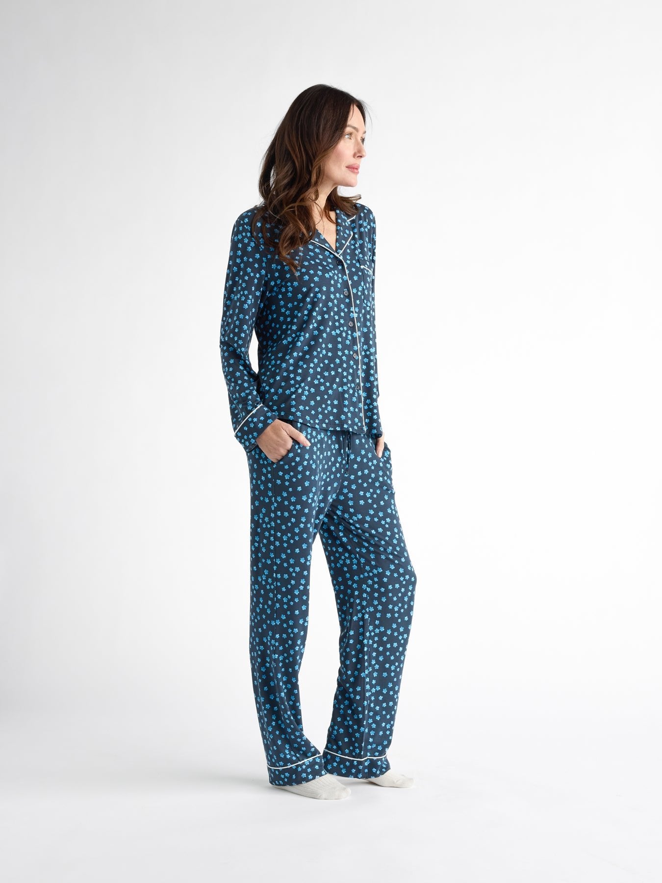 A person stands against a white background, wearing Cozy Earth Women's Bamboo Stretch-Knit Long Sleeve Pajama Top in navy blue with white polka dots. They look to the side with hands in their pockets, completing the set with full-length pants.