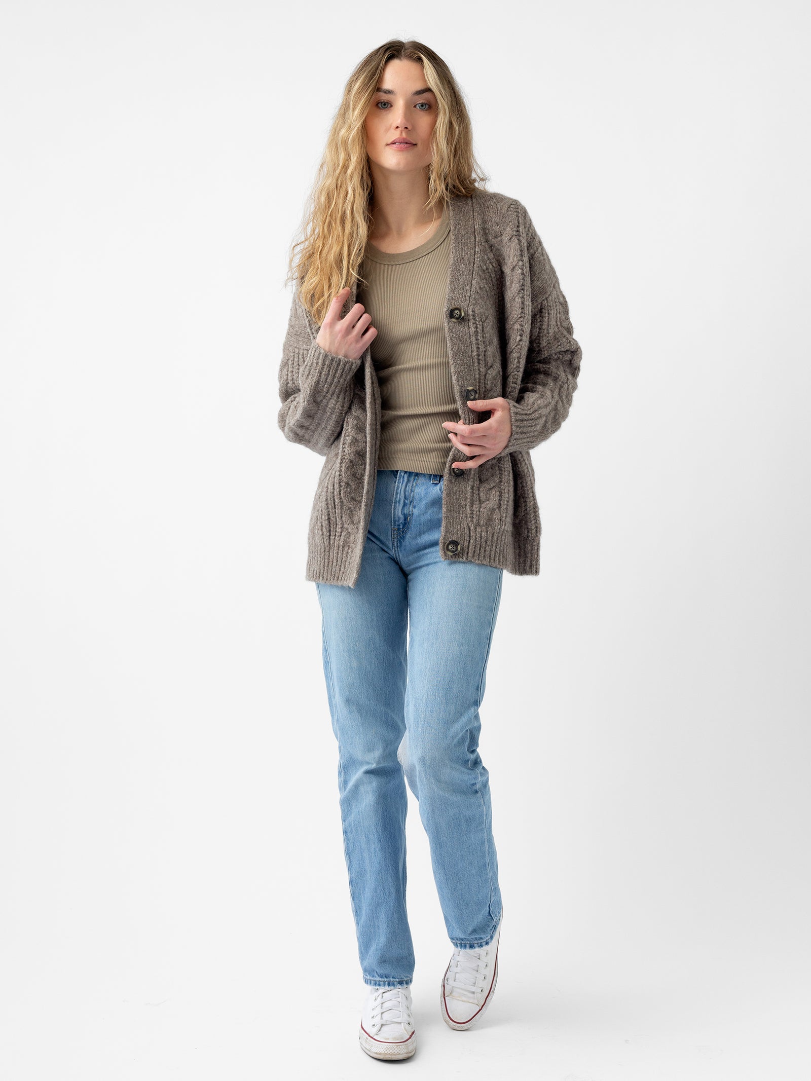 A person with long, wavy hair wears a beige top, light blue jeans, and white sneakers. They stand against a plain white background, gazing directly at the camera with one hand on their Cozy Earth Oversized Cable Knit Cardigan.