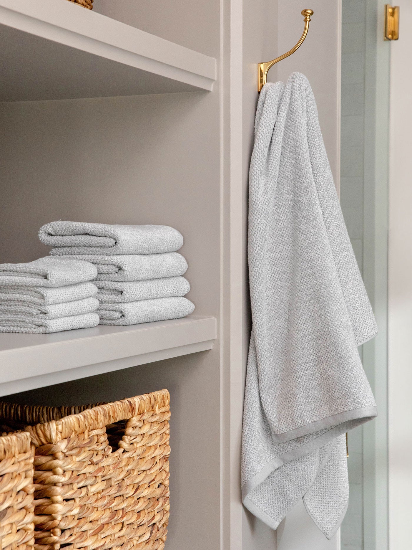 The Cozy Earth Complete Nantucket Bath Bundle features neatly stacked light gray towels on a shelf beside woven storage baskets, with two gray towels hanging on a gold hook in the bathroom.