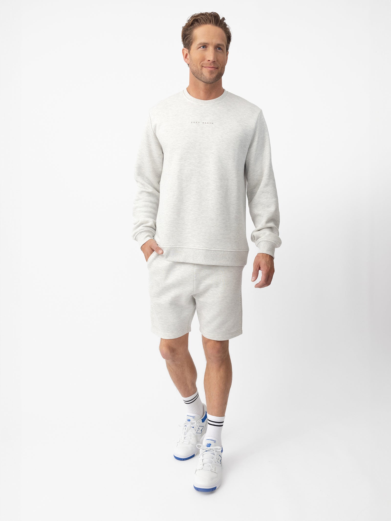 A man with short brown hair wears Cozy Earth’s Men's CityScape Shorts and a light gray sweatshirt, along with white socks and sneakers. He stands against a plain white background, looking slightly to the side and smiling.