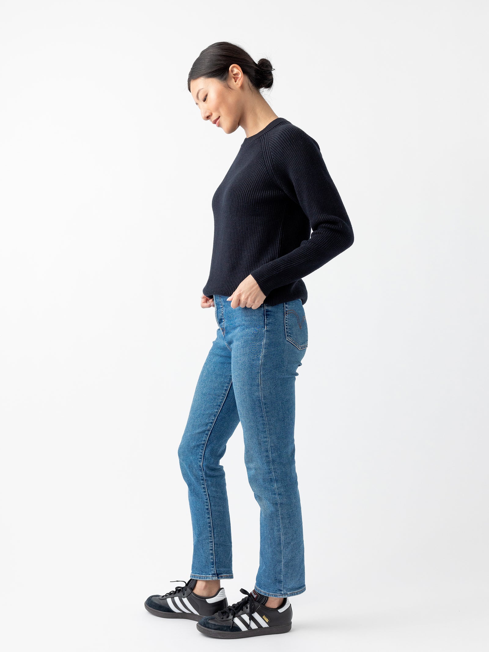 A woman wearing a Cozy Earth Women's Classic Crewneck, blue jeans, and black sneakers with white stripes stands against a plain white background. She is facing slightly to the left with her hands in her pockets and her hair tied back.
