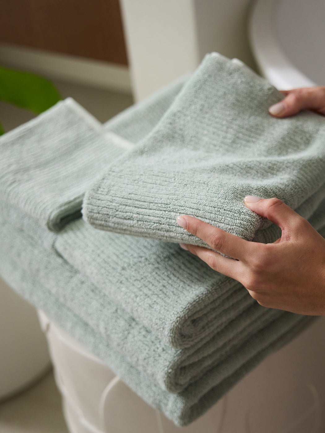 A person holds a soft, light green Complete Ribbed Terry Bath Bundle by Cozy Earth, with two more folded towels stacked below in a bathroom setting.