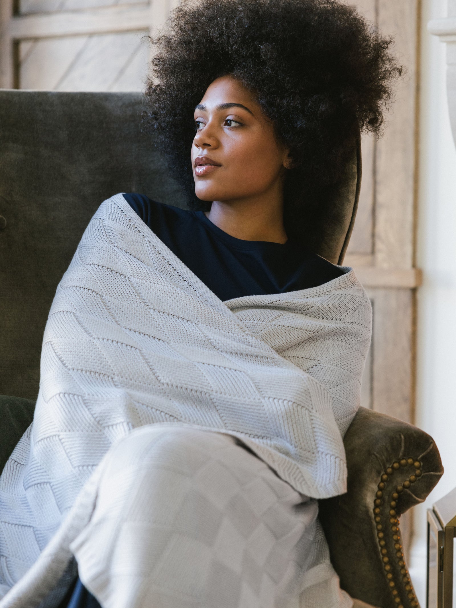 Light Grey Diamond Knit Blanket photographed in a home living room. A woman holds the blanket over her shoulder. 