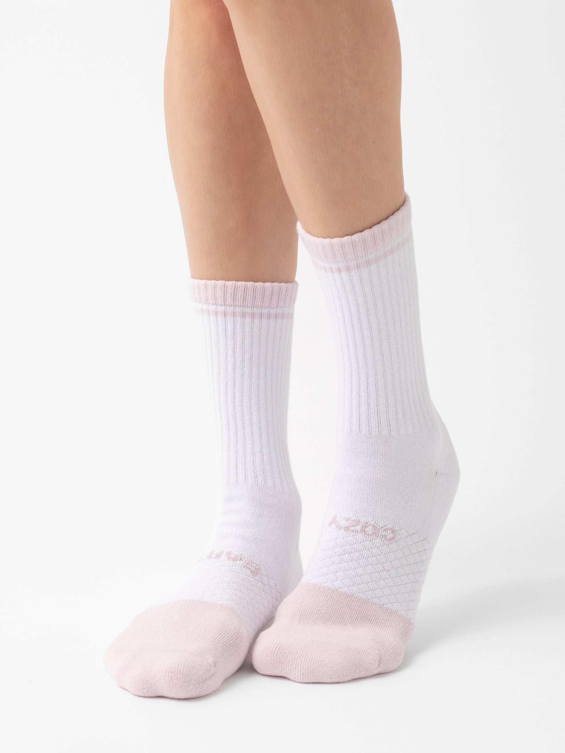 Close-up of a pair of legs wearing white Essential Calf Socks from Cozy Earth, featuring light pink toe and heel accents. The socks have a ribbed texture around the ankles and the word "COZY" written near the toes. The background is plain white. |Color:Lilac