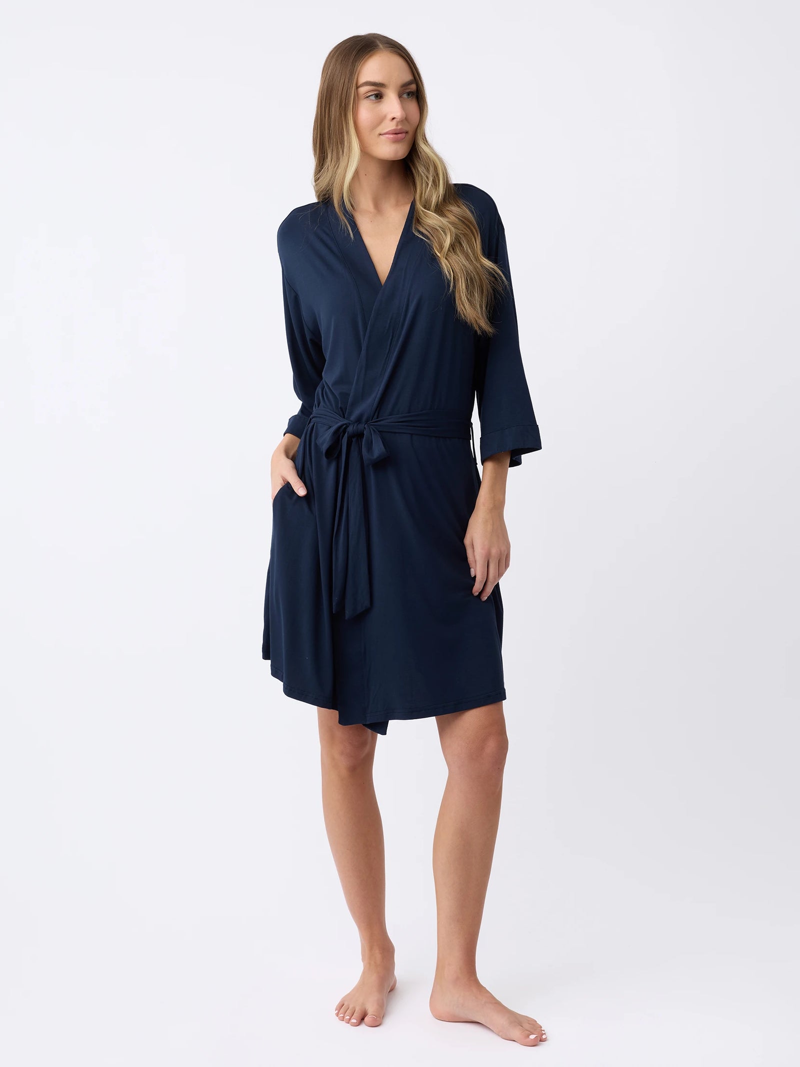A person with long hair poses barefoot against a plain white background, wearing Cozy Earth's Women's Stretch-Knit Bamboo Kimono Robe in navy blue, featuring long sleeves, a wrap style, and a waist tie.