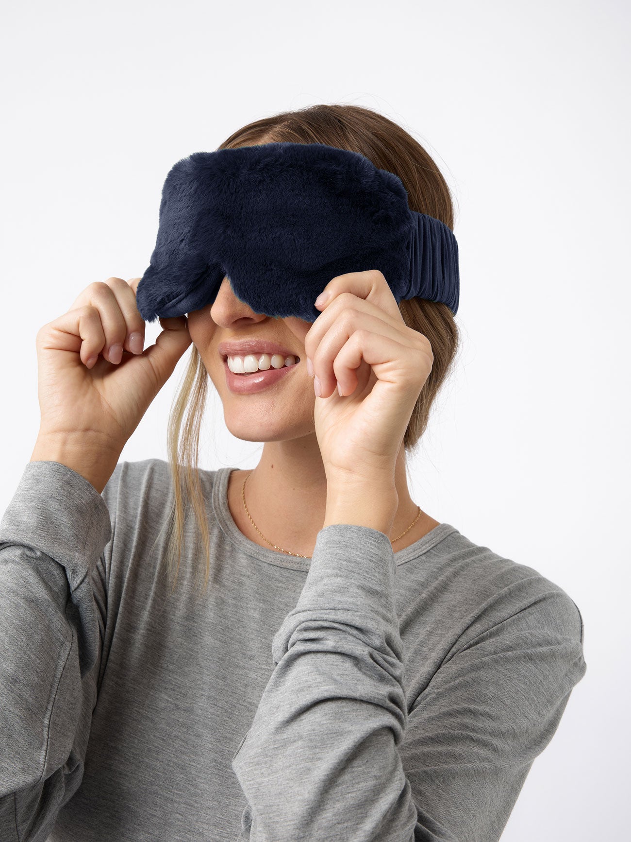 Someone in a gray long-sleeve shirt smiles as they adjust their Cozy Earth Faux Fur Sleep Mask, set against a plain white background.