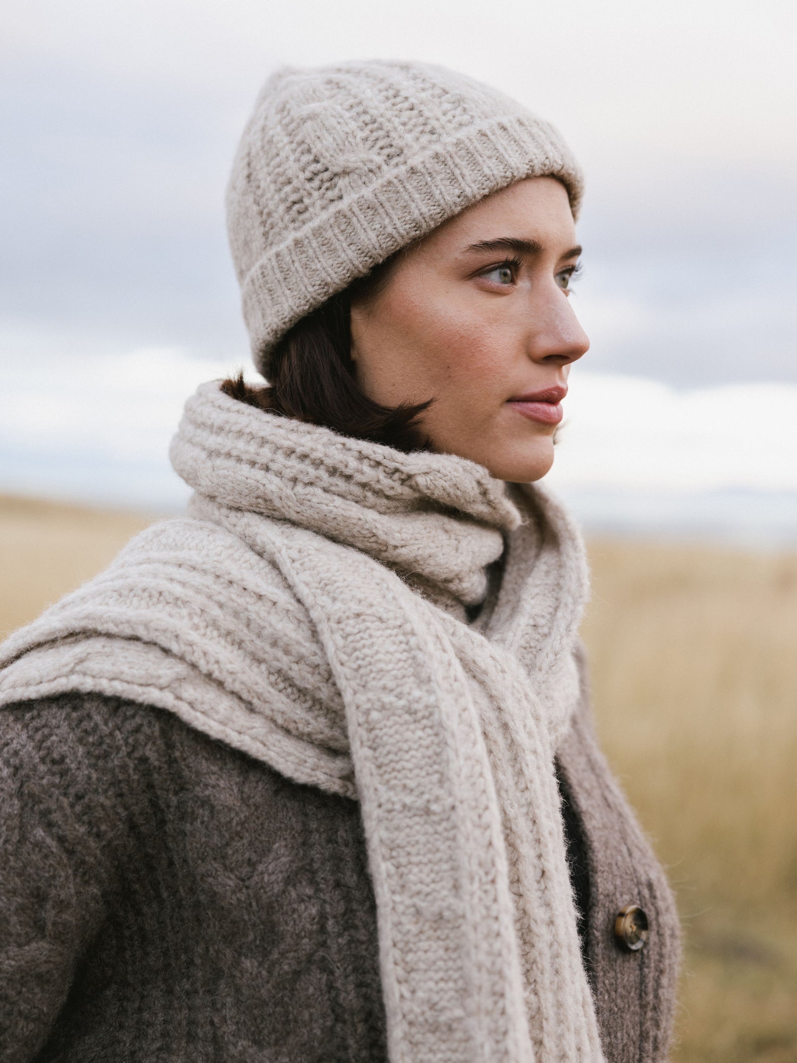 Woman wearing a tan Cozy Earth Cable Knit Beanie and scarf in a field of tall grass.